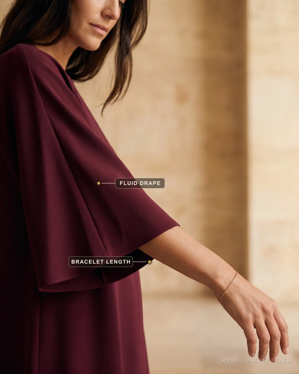 Fluid three-quarter sleeve drape in matte crepe for over 50 | Hugo Caesar Tassi Close detail of a deep-wine matte-crepe three-quarter sleeve, annotated with a gilt dot on the fluid drape and on the bracelet-length hem above a bare forearm with a fine gold bracelet.