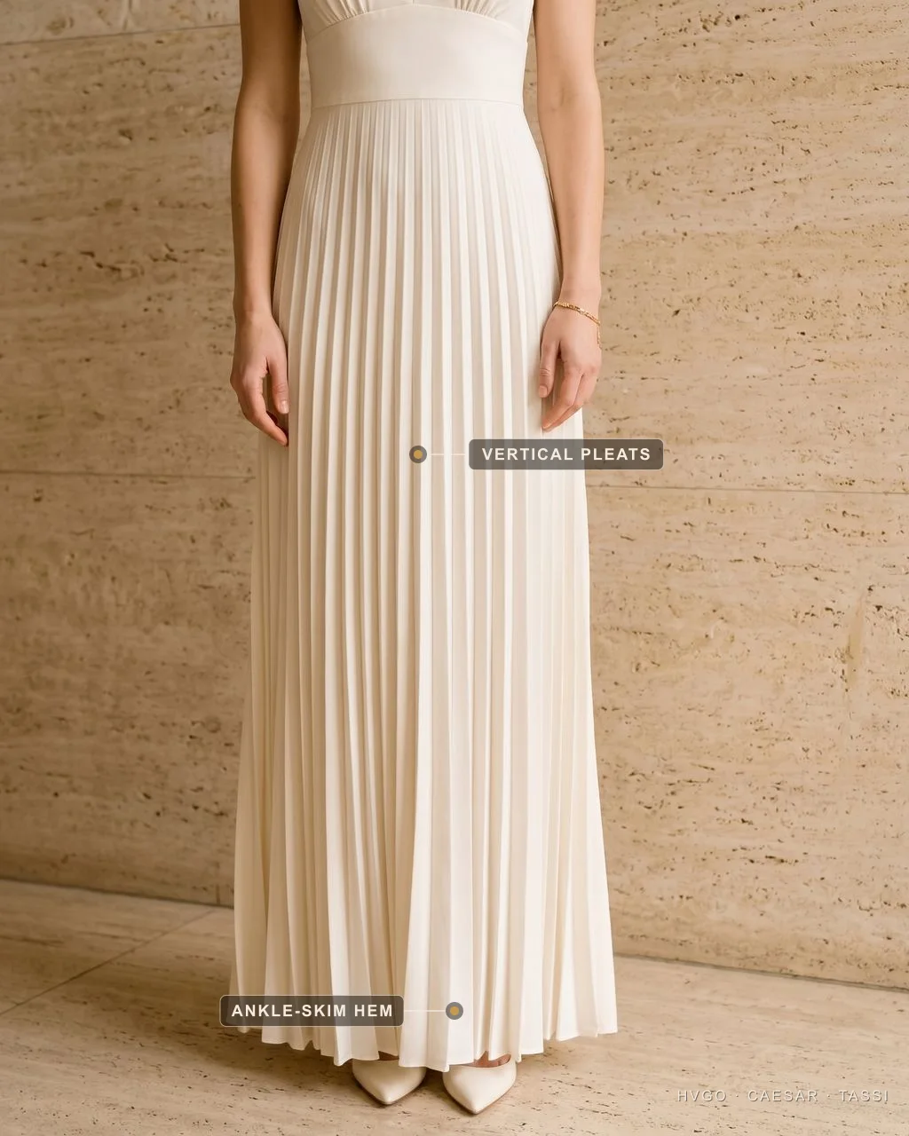 Vertical pleats from a high seam to an ankle-skim hem on a petite gown — Hugo Caesar Tassi A close detail of an Avorio ivory pleated column gown, no face in frame, with a label on the fine vertical knife pleats falling from a high empire seam and a second label at the ankle-skimming hem above pointed pumps.