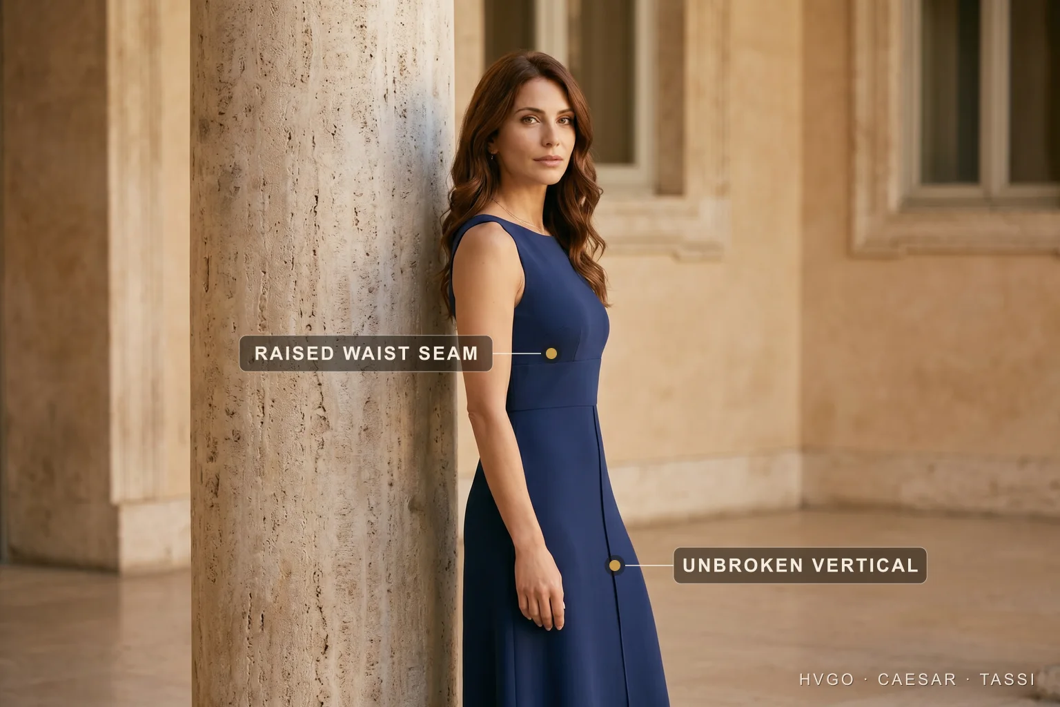 One-colour navy column with a raised waist seam for a petite frame — Hugo Caesar Tassi Sofia in a Cobalto navy one-colour column gown leaning against a travertine column, with a label marking the raised waist seam at her midriff and a second marking the unbroken vertical line running down the centre-front of the skirt.