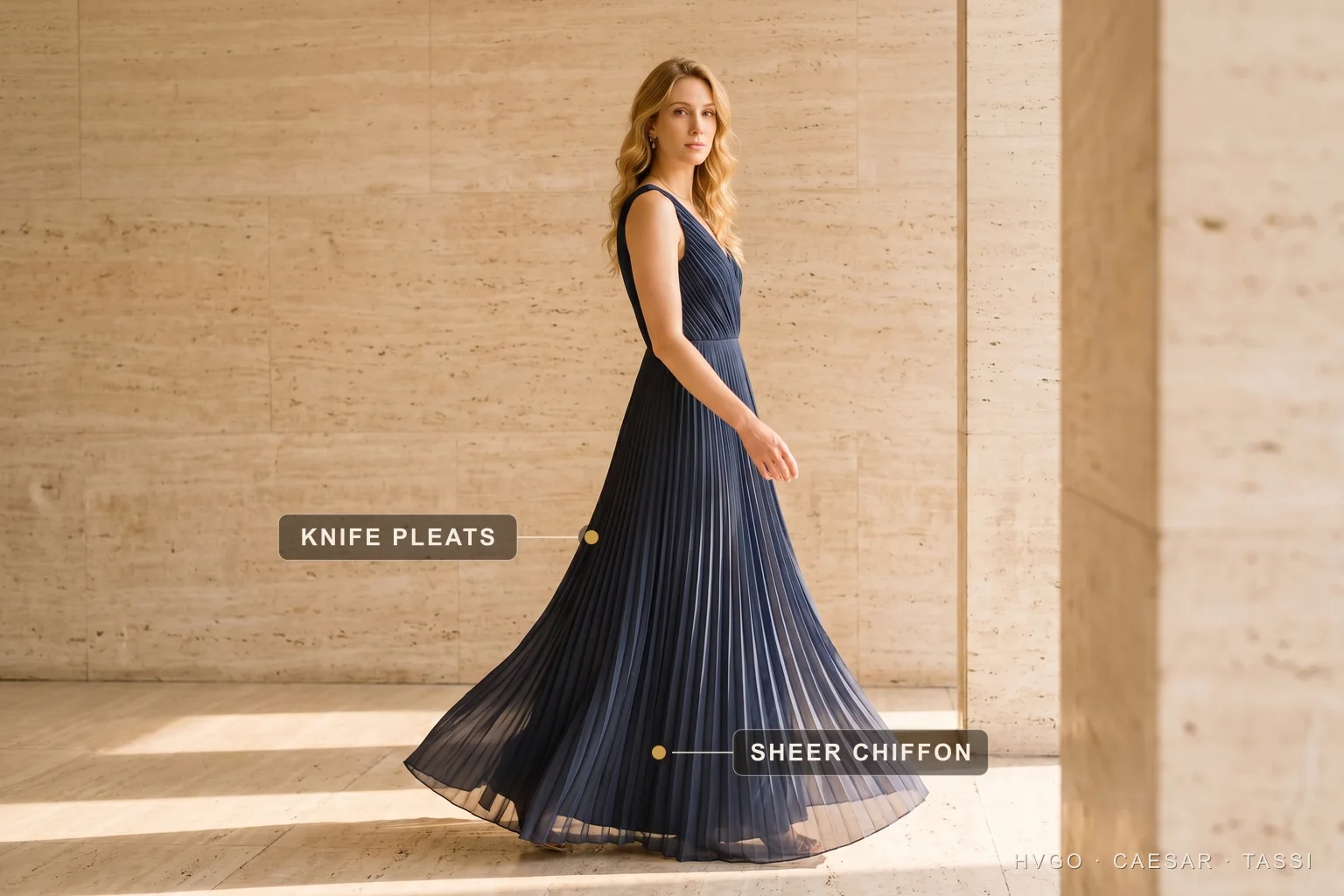 Knife-pleated chiffon gown catching the light — Hugo Caesar Tassi A model in a floor-length navy pleated chiffon gown in a sunlit travertine interior, with a gilt marker on the skirt's vertical folds labelled Knife pleats and a second marker on the translucent lower hem labelled Sheer chiffon.