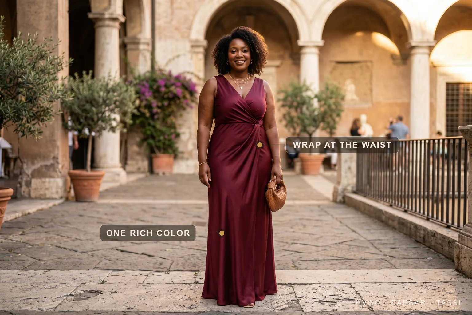 Plus-size wrap occasion gown in wine — Hugo Caesar Tassi A confident plus-size woman in a Hugo Caesar Tassi wine bias-cut satin wrap gown in a sunlit Roman courtyard, labelled to show the wrap crossing and defining the waist and one saturated color running in a long line