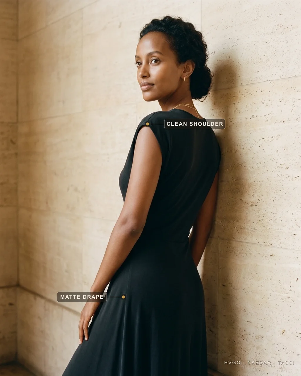 Quiet luxury tells — the clean shoulder seam and matte drape on a black modal gown — Hugo Caesar Tassi Aisha in a Hugo Caesar Tassi Basalt-black matte modal gown shown in three-quarter-back profile glancing over her shoulder against a plain Roman travertine wall, labelled to show the shoulder seam sitting exactly at the shoulder edge and the matte drape falling from the hip