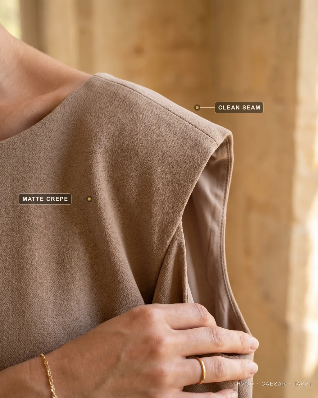 Extreme close-up of a camel-stone occasion gown's shoulder, with baked labels marking the clean shoulder seam running precisely along the shoulder line and the matte crepe fabric surface, a hand with a fine gold ring resting against the cloth and no visible hardware.