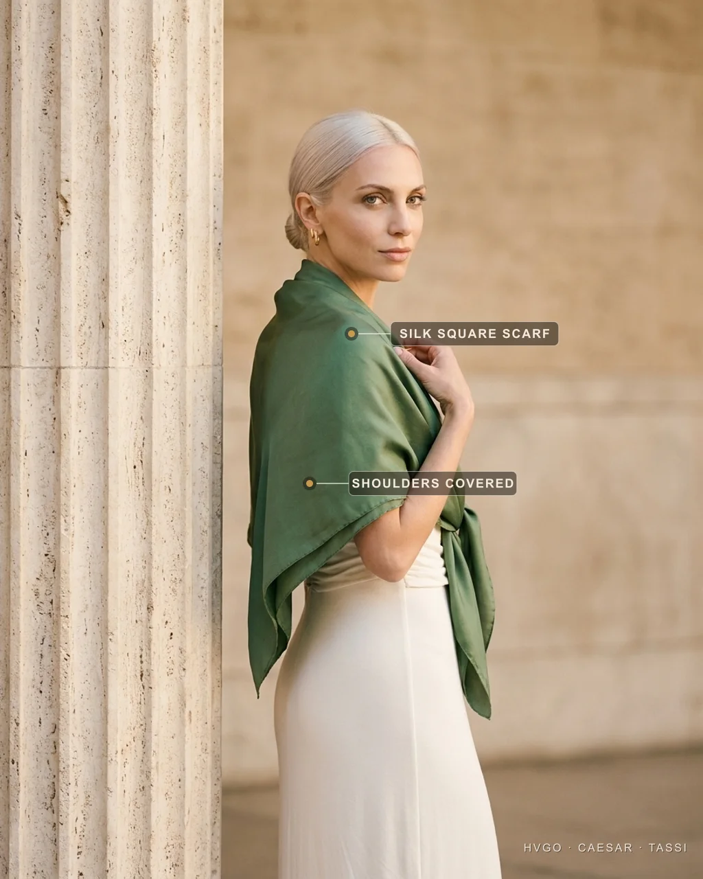 Valentina in three-quarter-back profile beside a fluted travertine column wearing a Hugo Caesar Tassi Avorio ivory draped modal column gown with a Verde Antico green silk square scarf draped over both shoulders, labelled to show the silk square scarf and the covered shoulders for a lakeside church