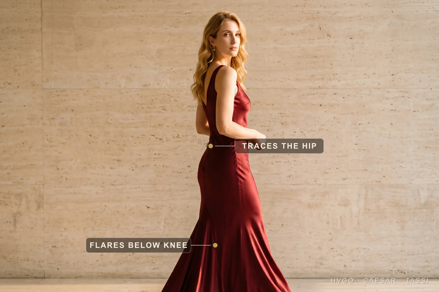 Dress silhouettes explained — the fishtail trumpet line — Hugo Caesar Tassi A golden-blonde woman in a Hugo Caesar Tassi Porpora deep-wine bias-cut satin fishtail gown shown in three-quarter-back profile in a Roman travertine studio, labelled to show the satin tracing the hip and the fishtail flaring out below the knee