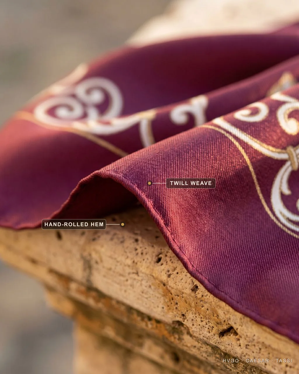 Macro close-up of a wine silk scarf with ivory-and-gold scrollwork print draped over a travertine ledge; labels mark the diagonal twill weave and the hand-rolled hem along the edge.