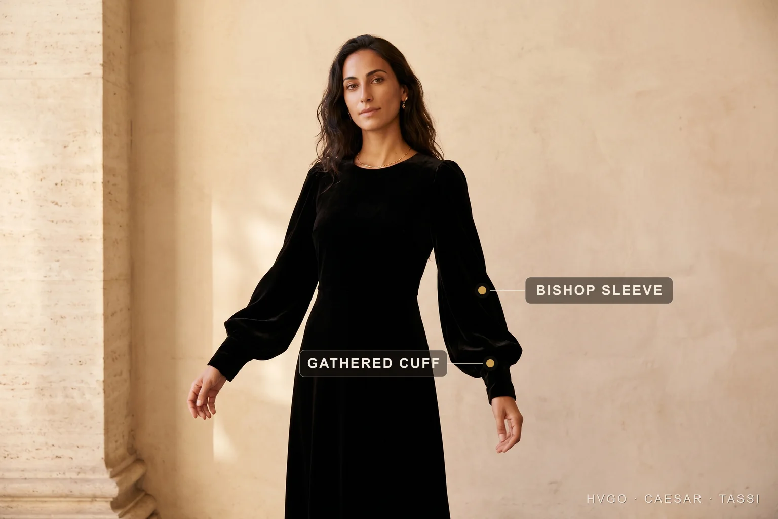 Dress sleeve types explained — the bishop sleeve — Hugo Caesar Tassi A woman in a Hugo Caesar Tassi black velvet floor-length gown with full-length bishop sleeves, in a Roman travertine setting, labelled to show the gathered bishop sleeve and the snug gathered cuff at the wrist