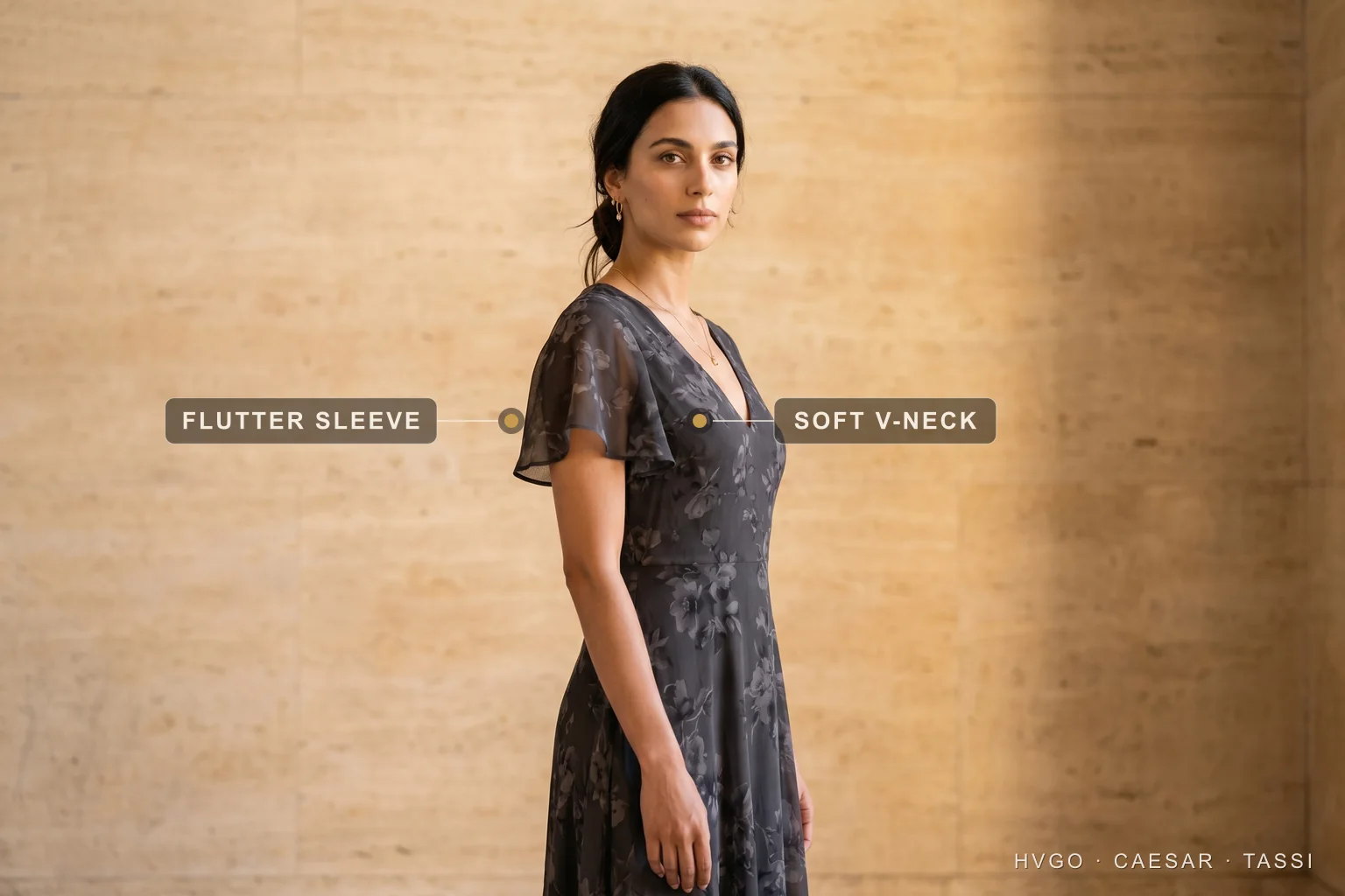 Dress sleeve types explained — the flutter sleeve — Hugo Caesar Tassi A woman in a Hugo Caesar Tassi charcoal floral-print chiffon A-line dress with a soft flutter sleeve and a soft V-neck, in a Roman travertine studio, labelled to show the flutter sleeve draping the upper arm and the soft V-neckline