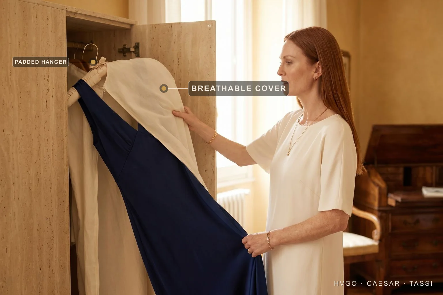 Storing an occasion dress on a padded hanger under a breathable cover — Hugo Caesar Tassi An auburn-haired woman beside an open travertine wardrobe holds a navy gown on a wide padded hanger, drawing back its ivory garment cover; labels mark the padded hanger and the breathable cover.