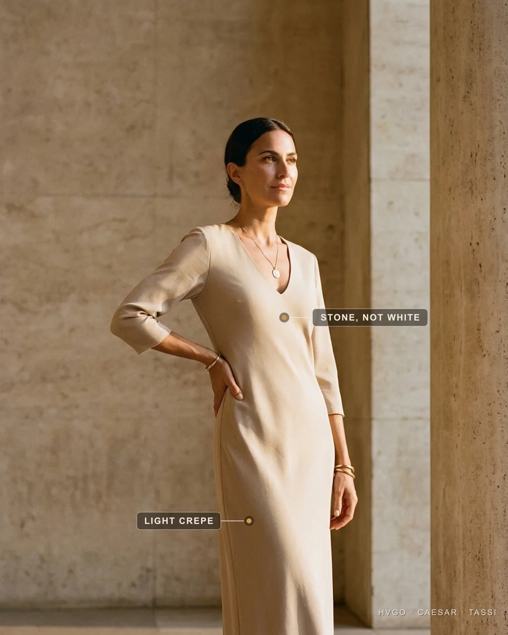 House model Marisol standing in a three-quarter contrapposto with one hand on her hip in a warm Travertine stone matte-crepe column dress with a soft V-neck and three-quarter sleeves, labelled to show that the stone tone is not white and that the cloth is light crepe, in a sunlit Roman travertine setting — a luminous daylight colour that never reads bridal