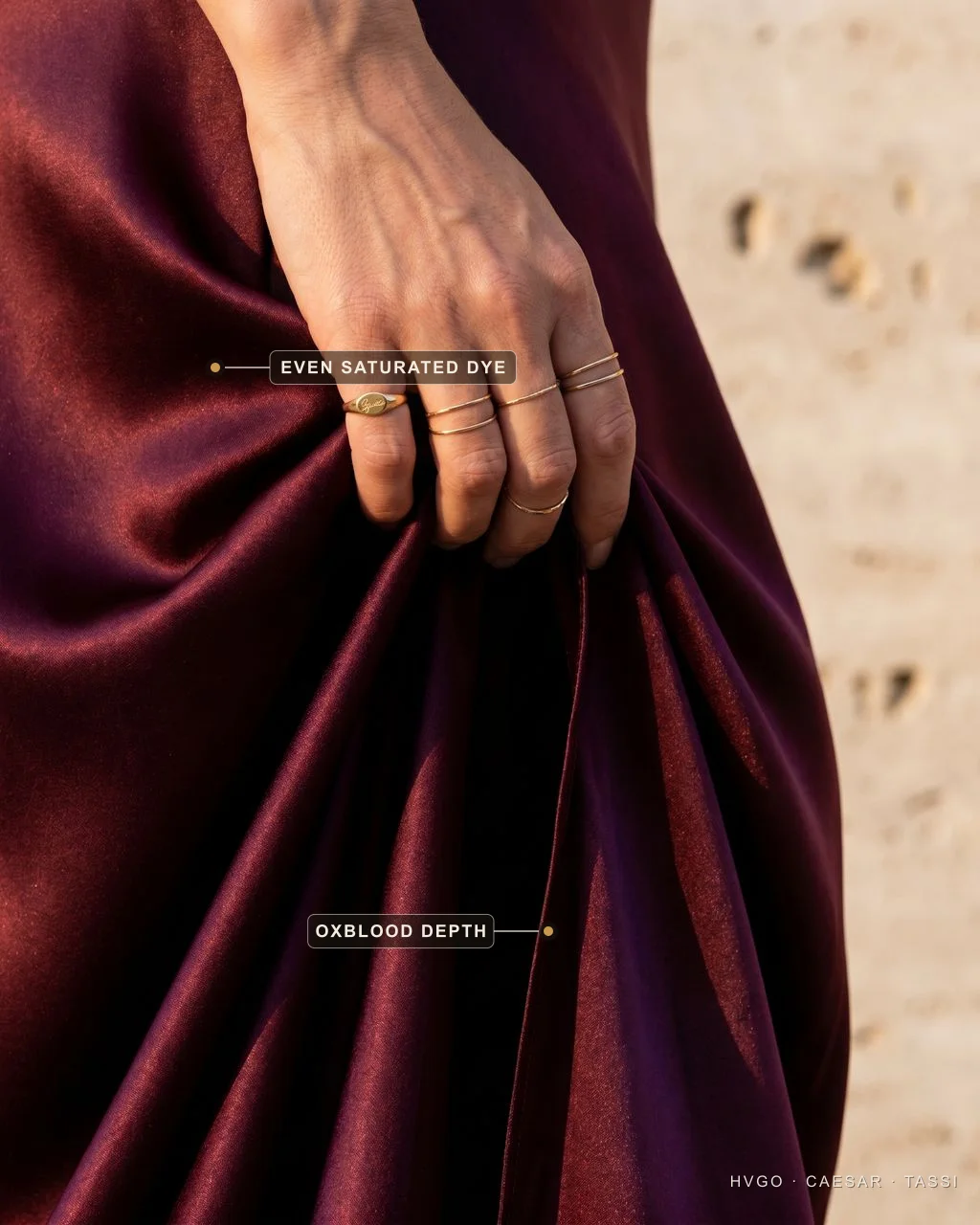 Even saturated oxblood dye depth in wine satin | Hugo Caesar Tassi Close fabric detail of a hand with fine gold rings lifting a fold of deep wine satin; baked labels mark the even saturated dye on the lit crest and the oxblood depth in the shadowed fold.