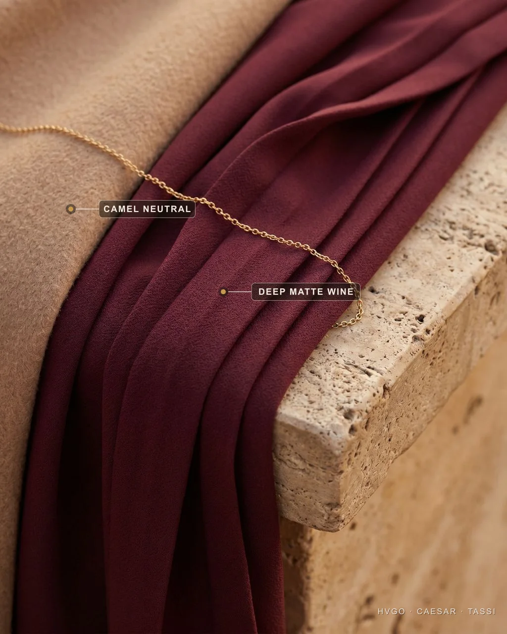 Matte wine crepe beside a camel neutral — Hugo Caesar Tassi Close-up of deep matte wine crepe fabric draped over a travertine ledge beside a soft camel cloth with a fine gold chain across the fold; labels mark the deep matte wine and the camel neutral.