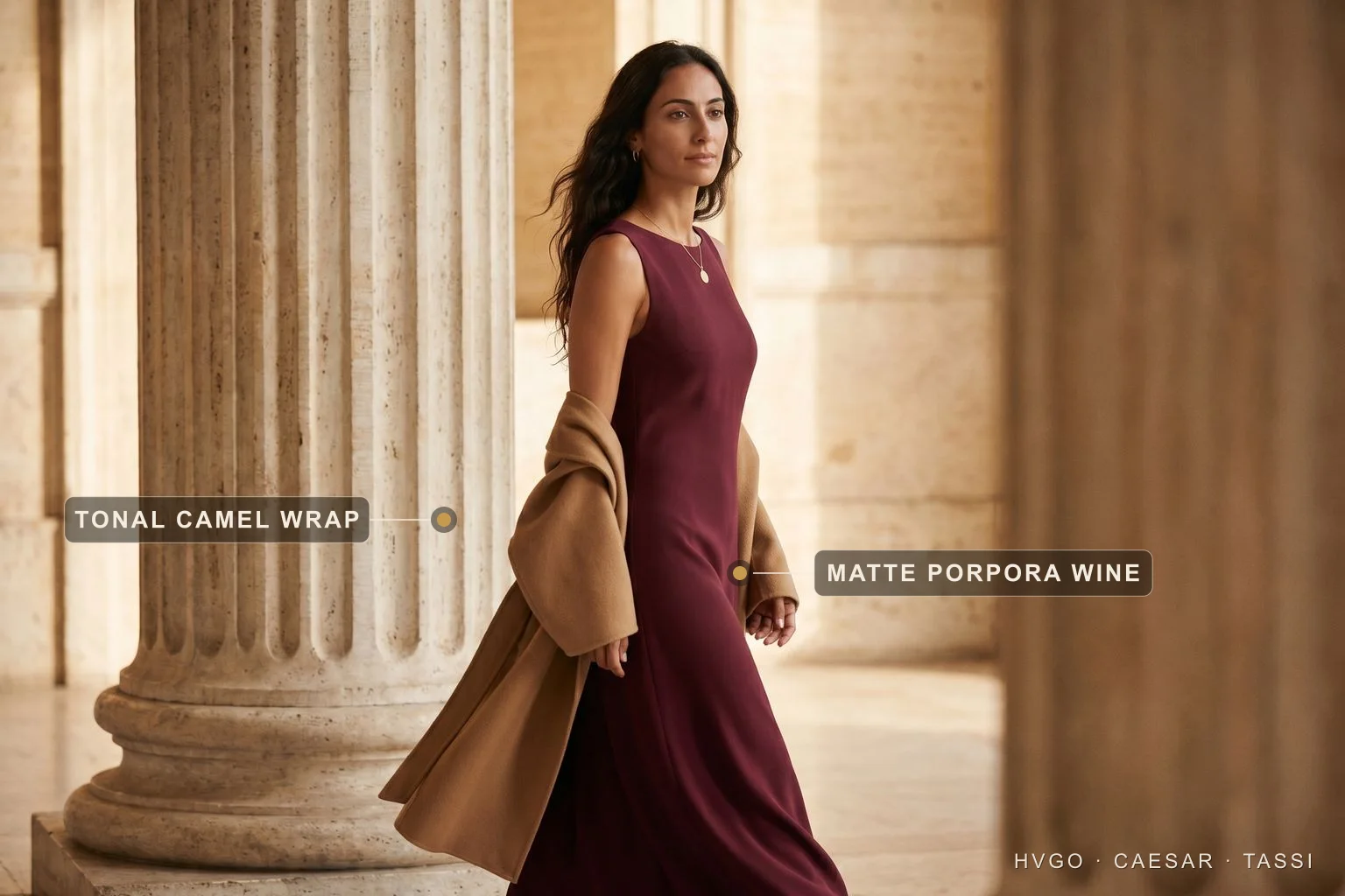 Burgundy dress styled tonally with camel — Hugo Caesar Tassi A dark-haired woman walking beside a travertine column in a deep matte Porpora wine column dress with a camel-stone wrap over her arm; labels mark the matte Porpora wine fabric and the tonal camel wrap.