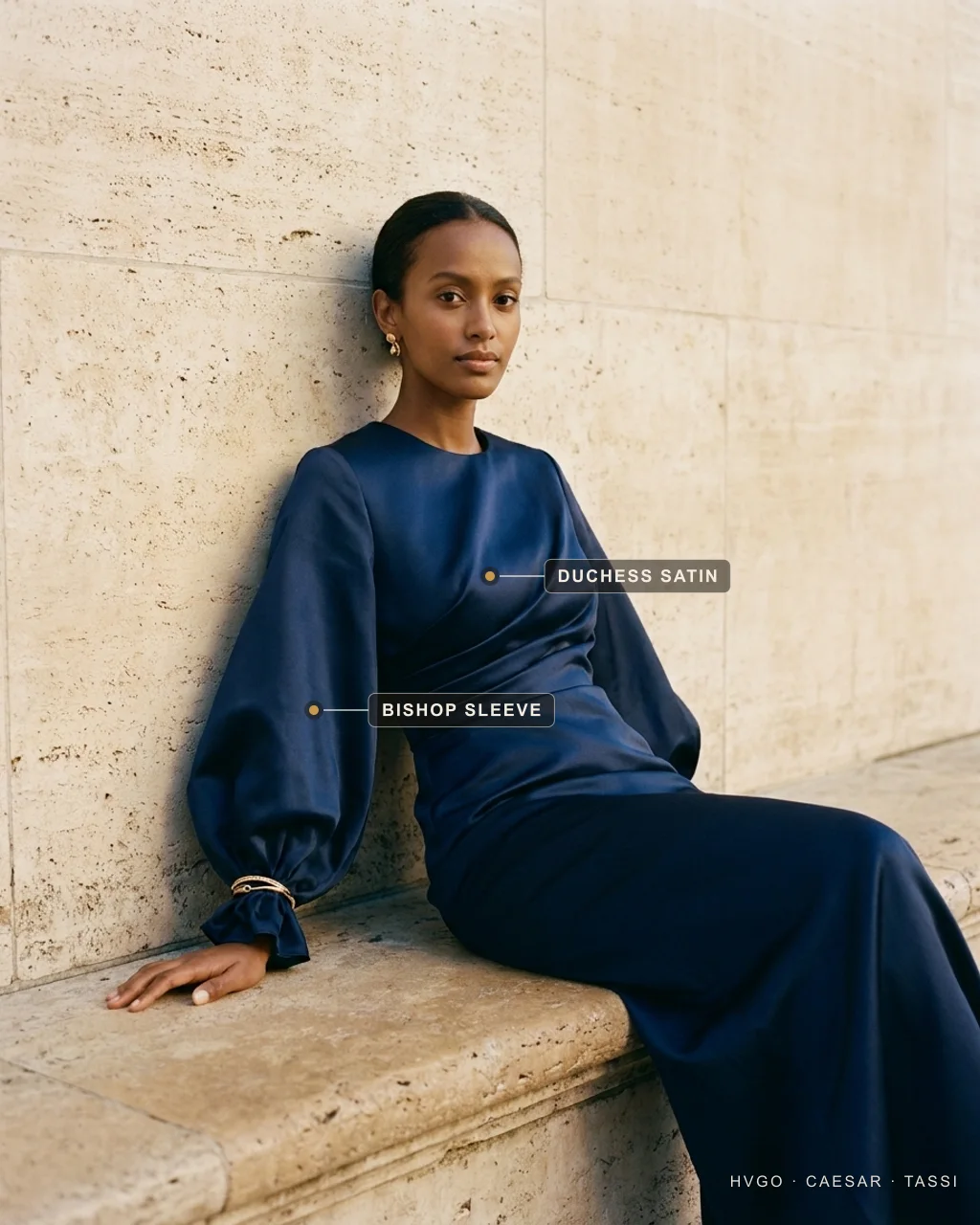 Navy Duchess-Satin Bishop-Sleeve Gown for a Winter Wedding Guest — Hugo Caesar Tassi Aisha seated and leaning on a travertine ledge in a deep navy duchess-satin gown with full bishop sleeves gathered at a gold-cuffed wrist, gilt markers labelling the duchess satin bodice and the bishop sleeve, in a warm Roman travertine setting