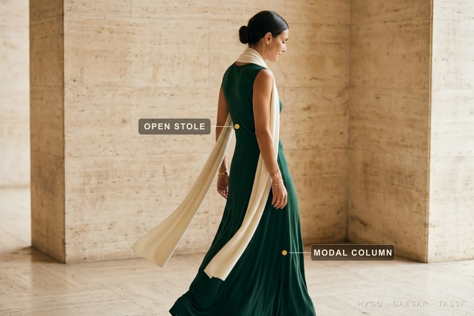 Emerald Modal Column with an Open Trailing Stole for a Winter Wedding Guest — Hugo Caesar Tassi Marisol walking mid-stride in three-quarter-back profile in a deep emerald modal-jersey column gown with an open ivory stole trailing long past the hip, gilt markers labelling the open stole and the modal column, in a warm Roman travertine setting