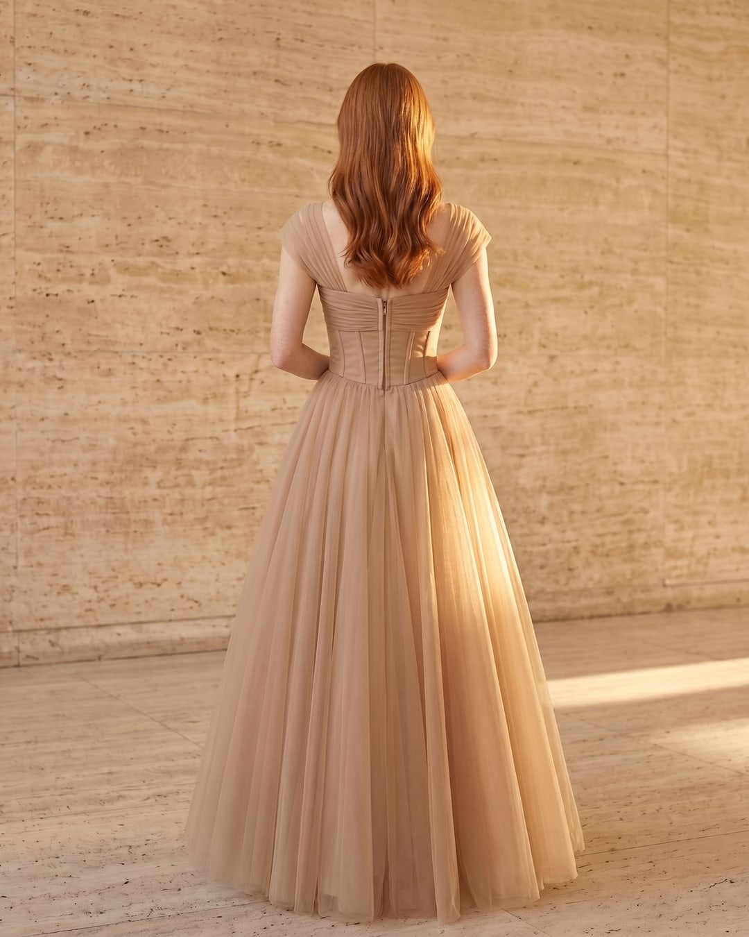Full-length rear view of the Hugo Caesar Tassi Julia French-style pleated chiffon maxi dress in Travertine apricot, seen from behind, showing the back of the dress, the soft surplice V-back, ruched waist and accordion-pleated floor-length skirt in a Roman travertine studio