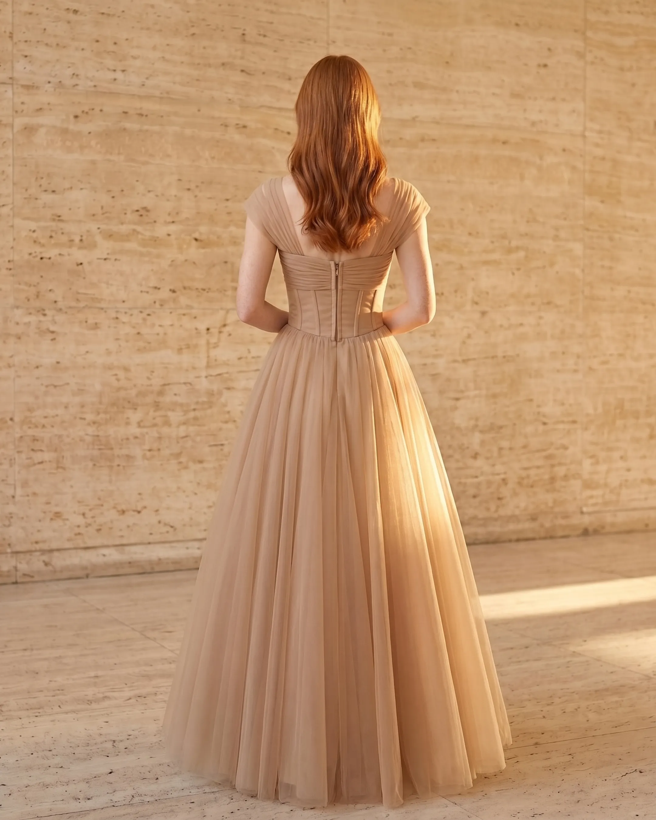 Full-length rear view of the Hugo Caesar Tassi Julia French-style pleated chiffon maxi dress in Travertine apricot, seen from behind, showing the back of the dress, the soft surplice V-back, ruched waist and accordion-pleated floor-length skirt in a Roman travertine studio