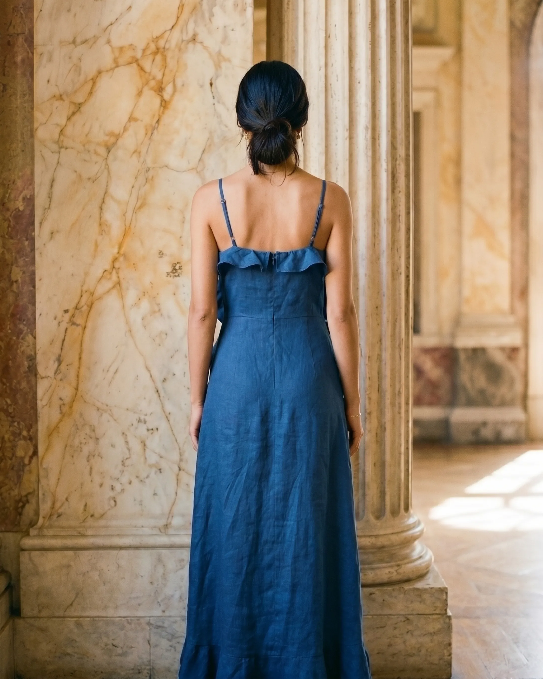 Full-length rear view of the Hugo Caesar Tassi Junia ruffle-trim slip midi dress in deep blue Cobalto linen-look crepe, seen from behind, showing the back of the dress, the fine straps, open back and easy midi slip line in a Roman travertine studio