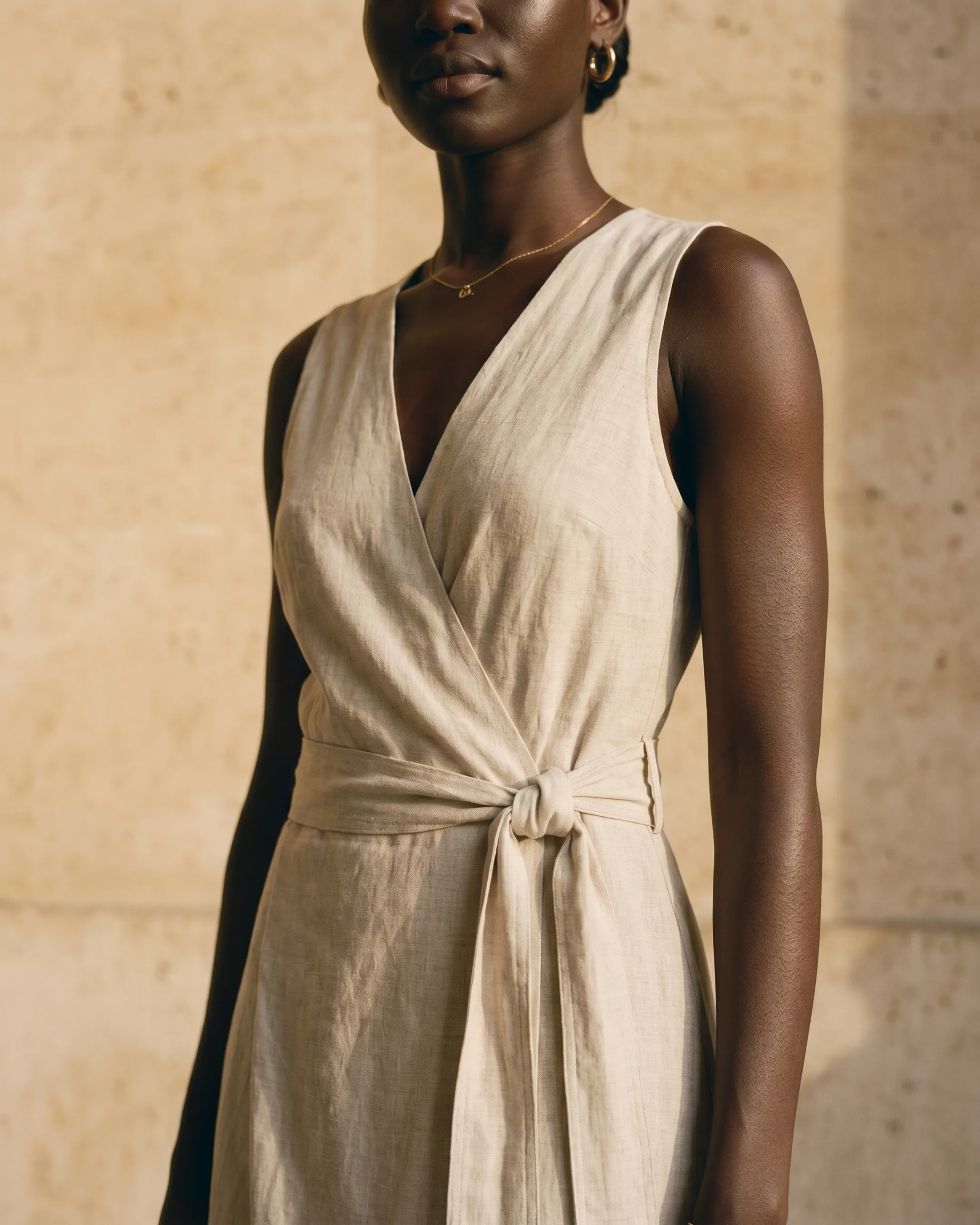 Detail of the Hugo Caesar Tassi Lavinia wrap dress in Avorio Tencel-linen, showing the deep V-neck faux-wrap bodice, sleeveless vest armhole and self-fabric tie knotted at the waist in a Roman travertine studio