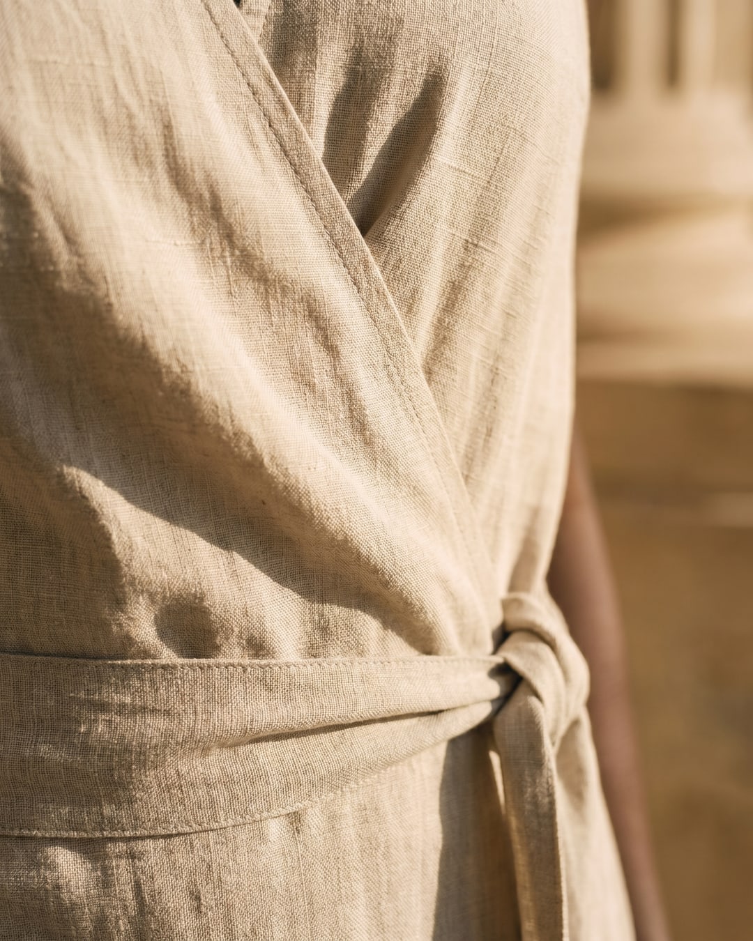 Macro detail of the Hugo Caesar Tassi Lavinia dress's matte, slubby Avorio Tencel-linen weave, the wrap overlap seam and self-fabric tie knot