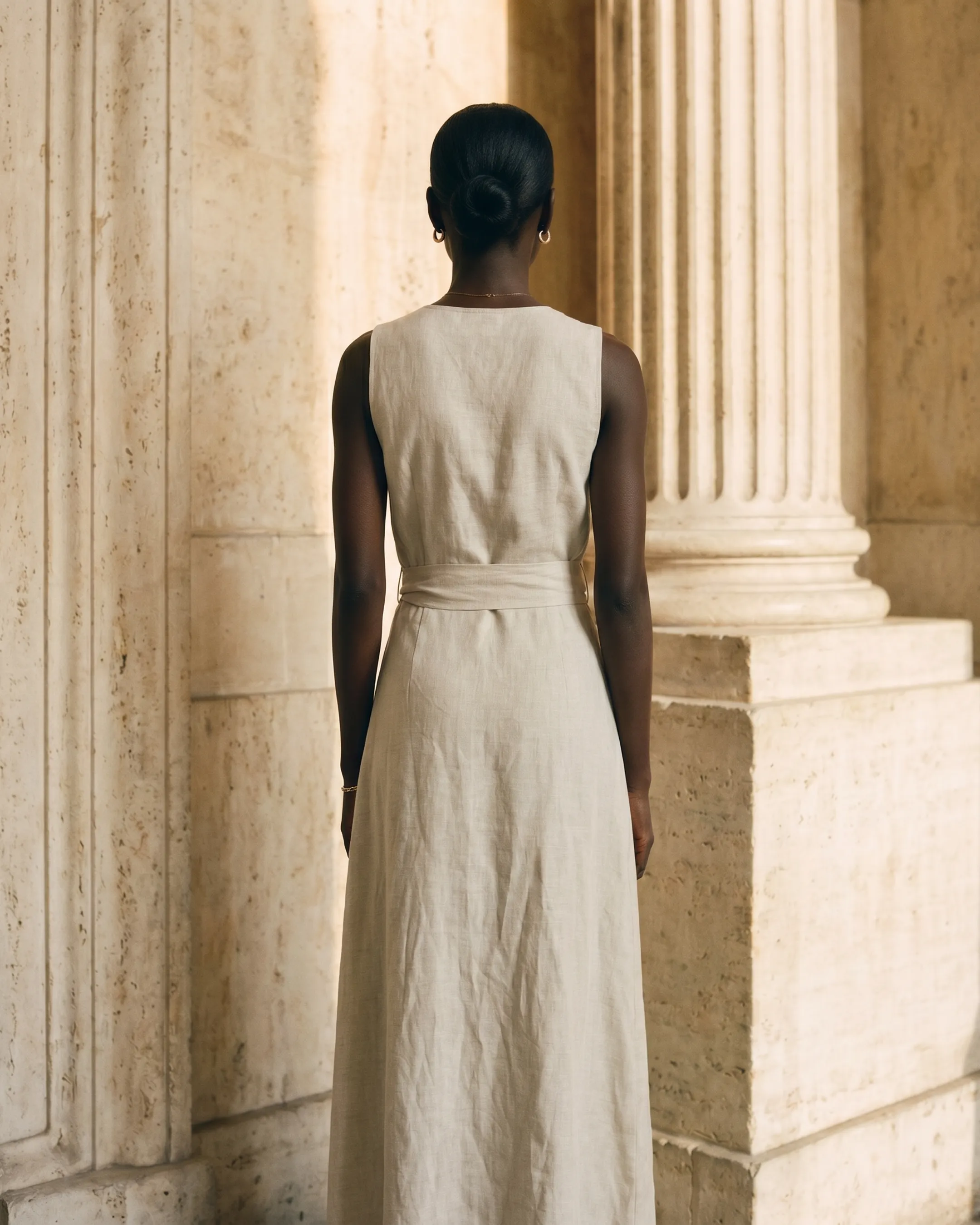 Full-length rear view of the Hugo Caesar Tassi Lavinia Tencel-linen wrap dress in Avorio, showing the clean back yoke, sleeveless vest armholes, wrapped self-tie sash and long A-line wrap skirt beside a Roman travertine column