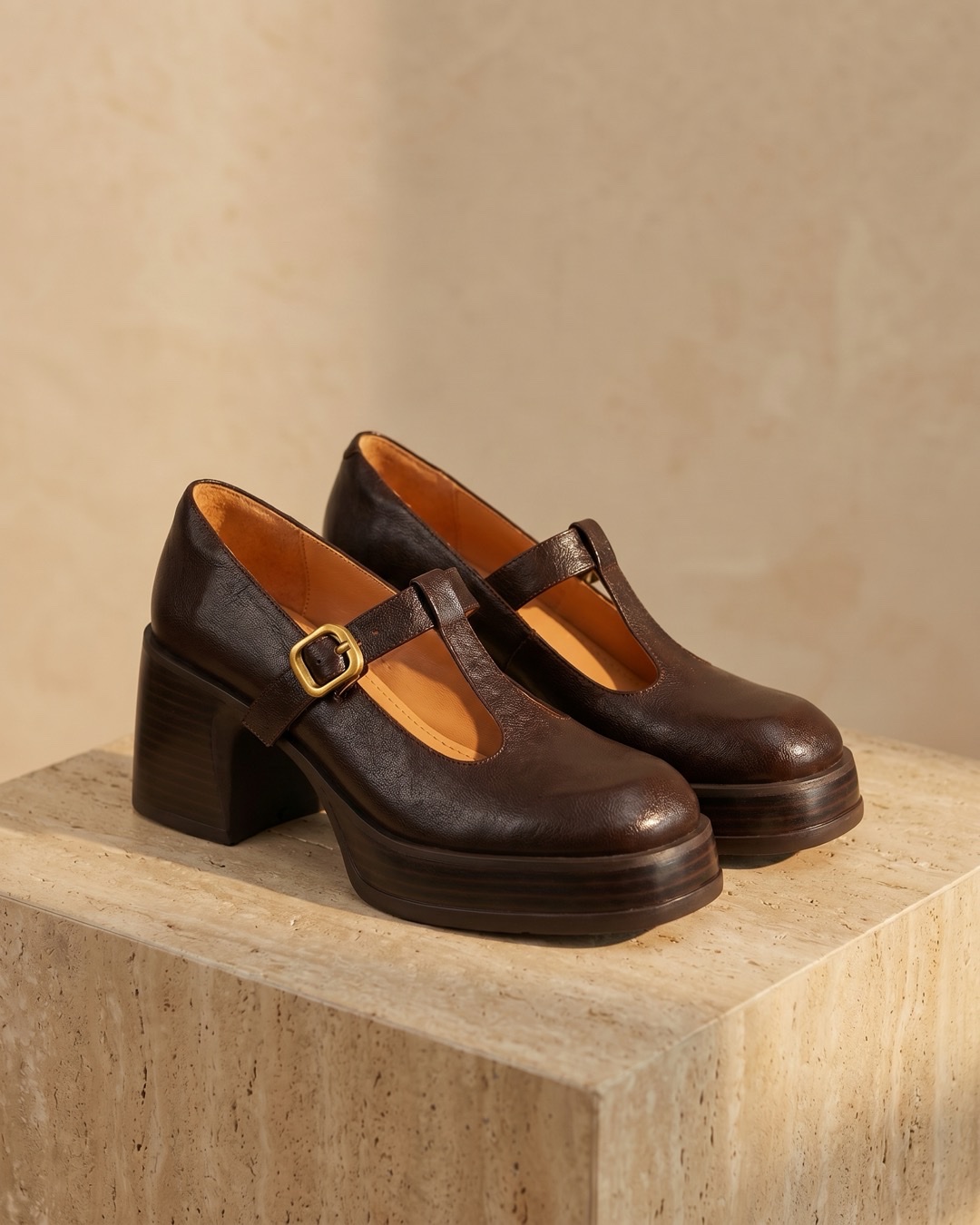 Hugo Caesar Tassi Lucilla T-bar block-heel leather Mary-Jane in Cuoio brown with a gold square buckle and platform sole, on a warm travertine pedestal
