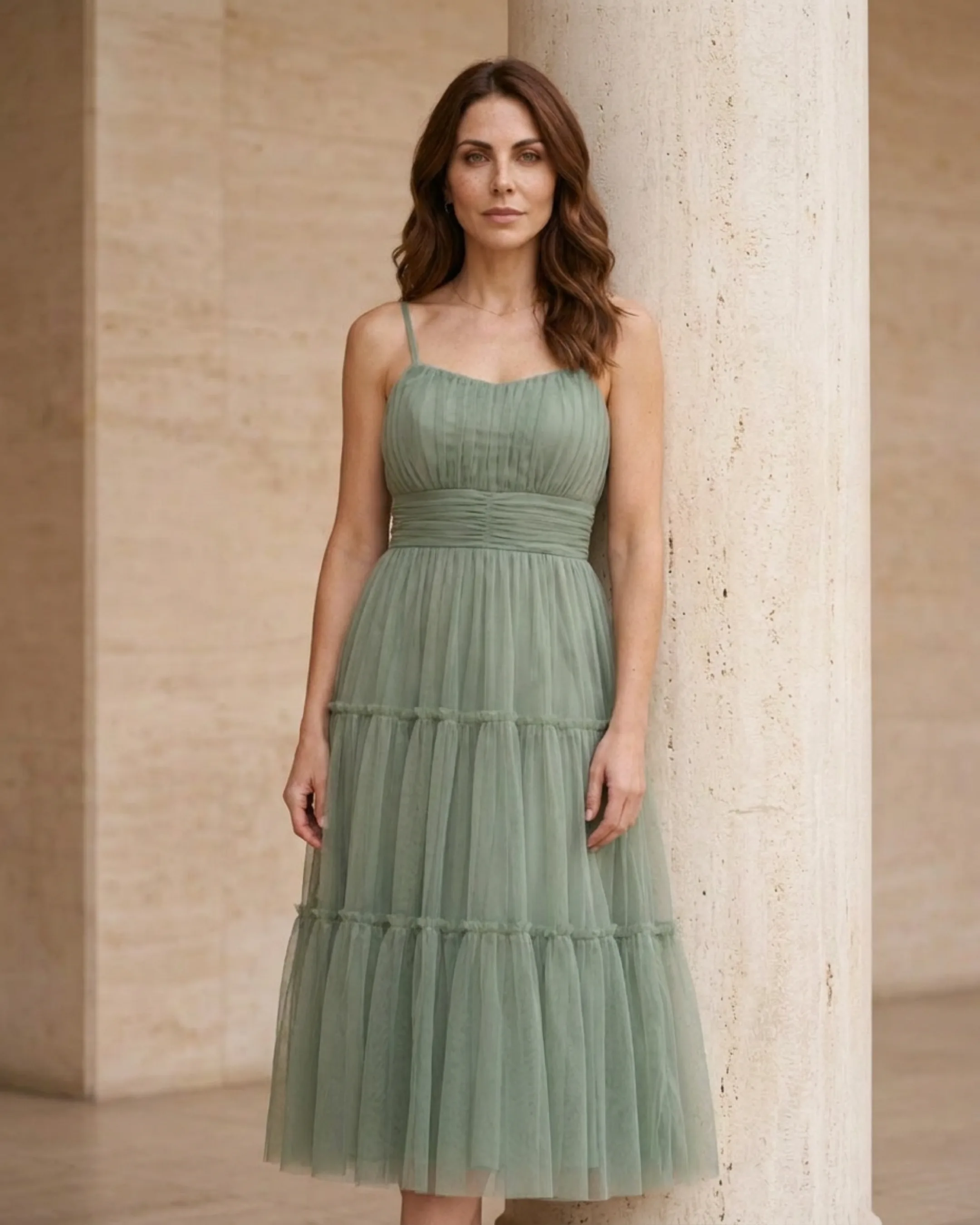 Hugo Caesar Tassi Marcia tiered tulle tea-length dress in soft sage green Verde Antico, worn by a model beside fluted columns in a Roman travertine studio