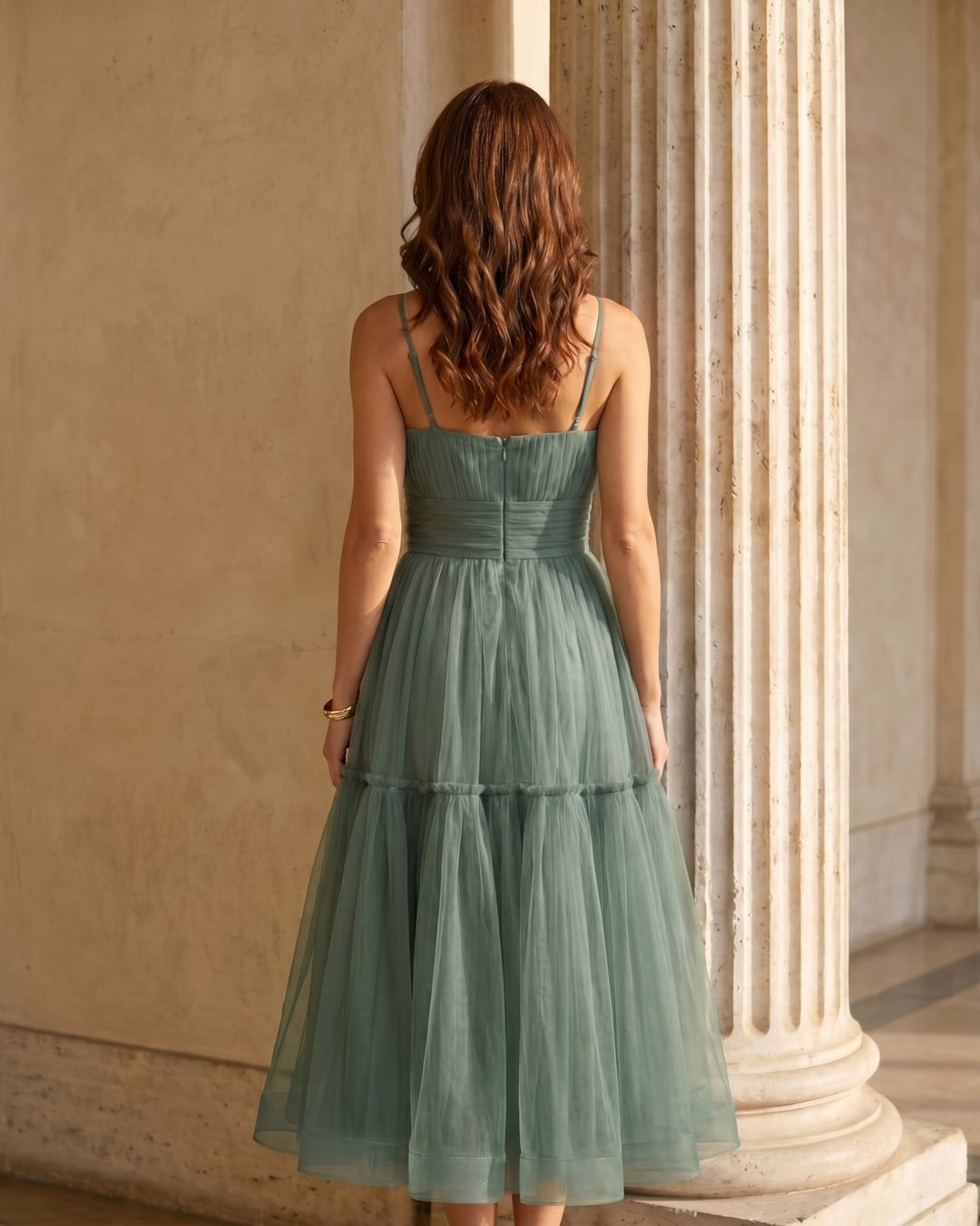 Full-length rear view of the Hugo Caesar Tassi Marcia tiered tulle tea-length dress in soft sage green Verde Antico, seen from behind, showing the back of the dress, the fine straps, lace-up corset back and full tiered tulle skirt in a Roman travertine studio
