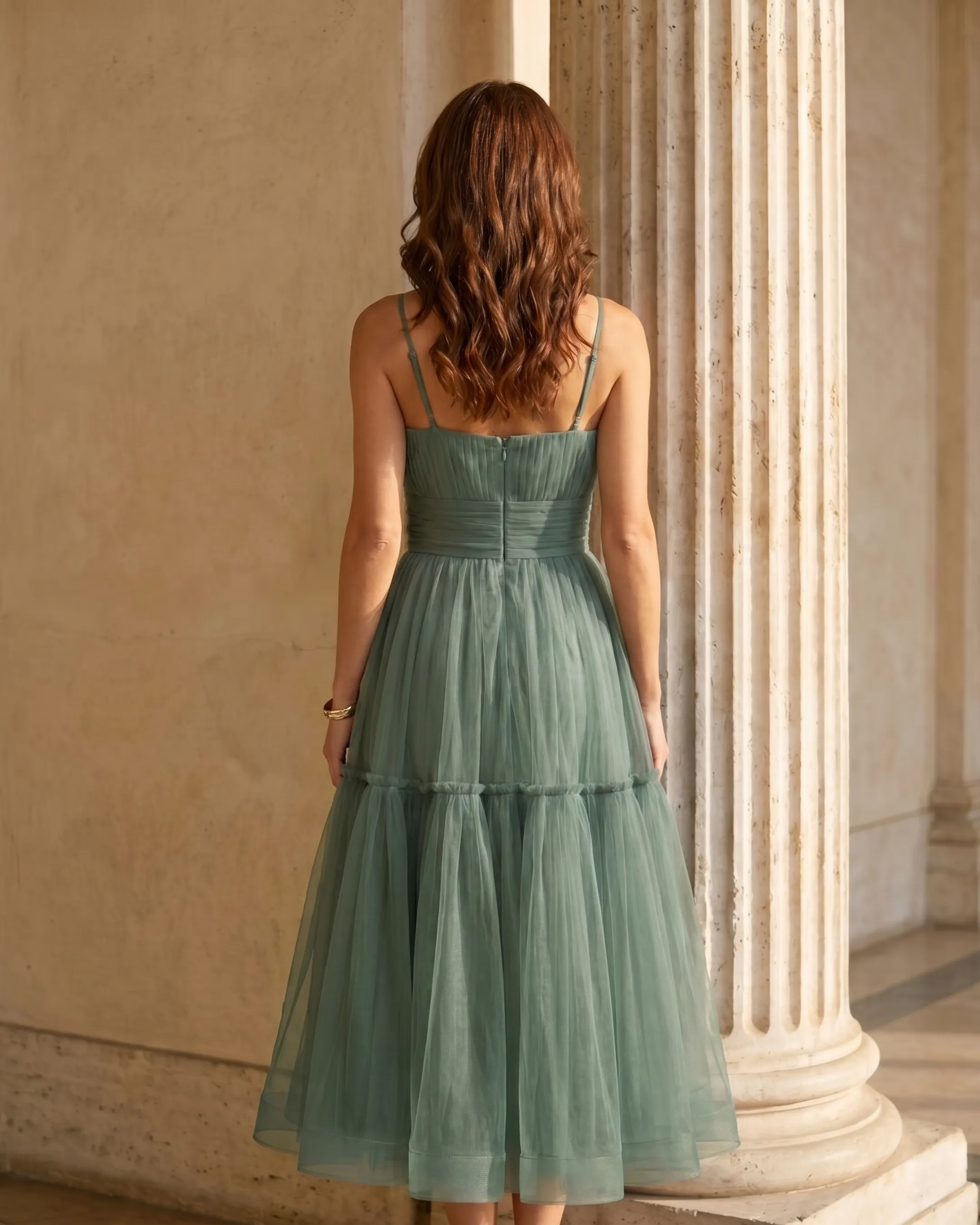 Full-length rear view of the Hugo Caesar Tassi Marcia tiered tulle tea-length dress in soft sage green Verde Antico, seen from behind, showing the back of the dress, the fine straps, lace-up corset back and full tiered tulle skirt in a Roman travertine studio