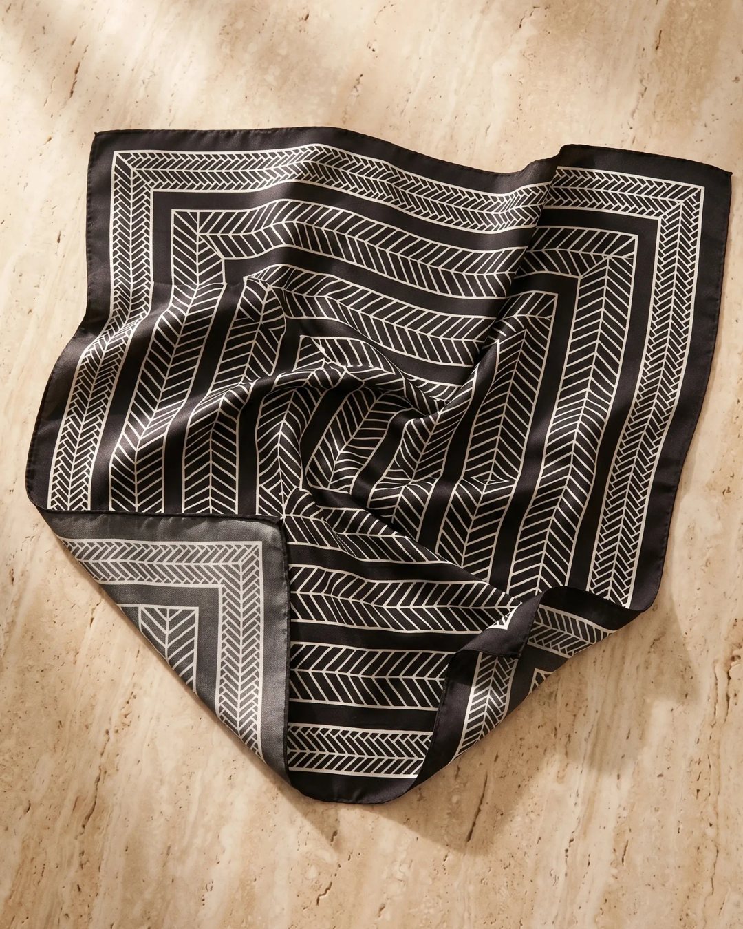 Hugo Caesar Tassi Palmyra scarf in Basalt — a styled flat-lay showing the full black-and-ivory concentric chevron print on a warm travertine surface