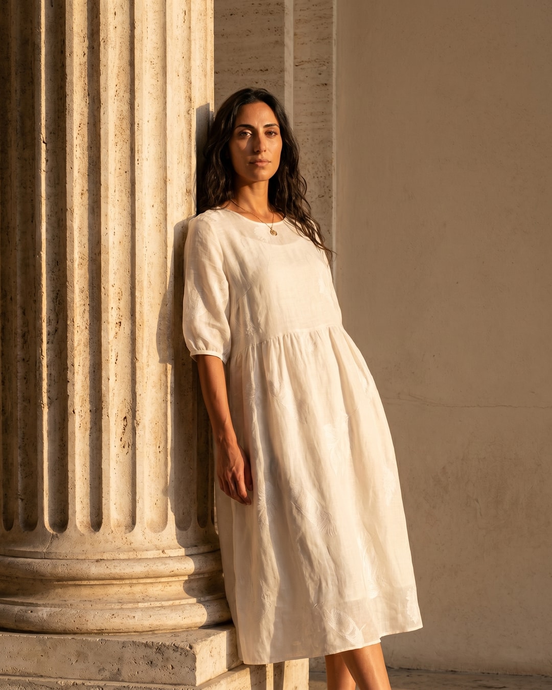 Editorial portrait of the Hugo Caesar Tassi Pomponia relaxed linen midi dress in Avorio, a model leaning on a fluted Roman travertine column in golden light