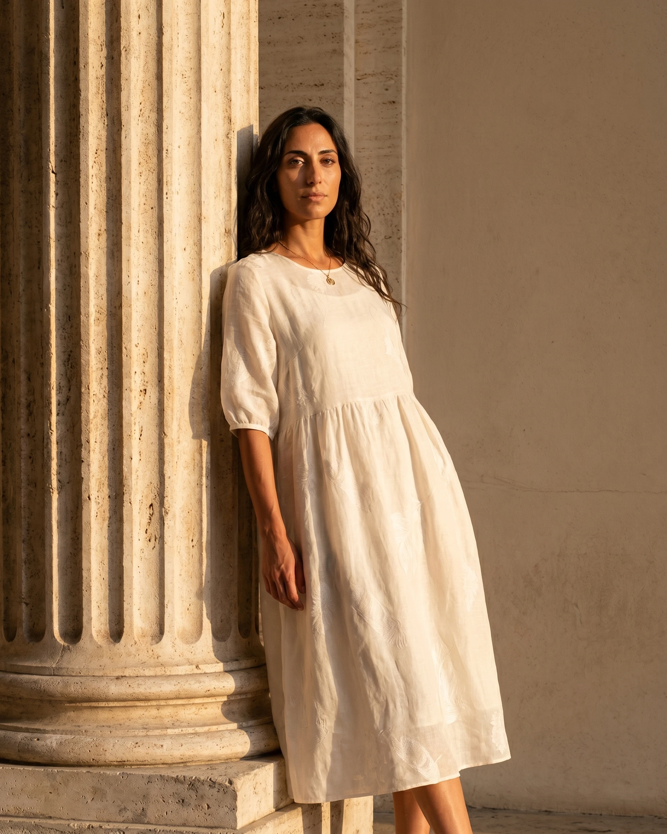Editorial portrait of the Hugo Caesar Tassi Pomponia relaxed linen midi dress in Avorio, a model leaning on a fluted Roman travertine column in golden light
