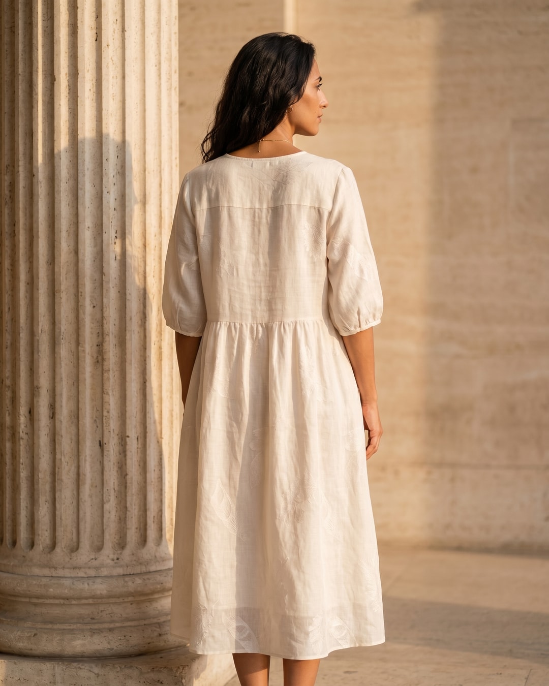 Full-length rear view of the Hugo Caesar Tassi Pomponia relaxed linen midi dress in Avorio ivory, showing the clean back yoke, drop-waist seam and easy A-line skirt beside a Roman travertine column