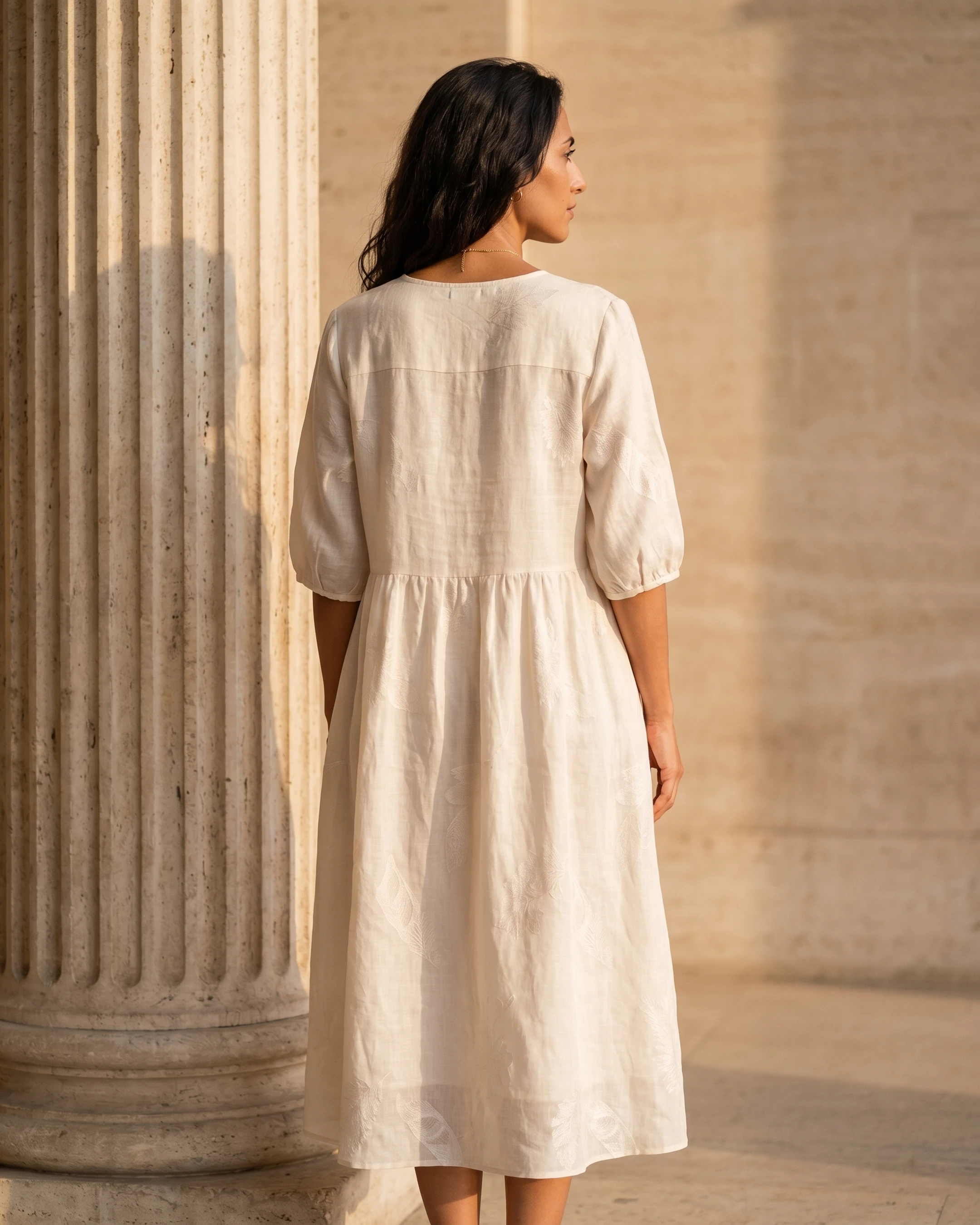 Full-length rear view of the Hugo Caesar Tassi Pomponia relaxed linen midi dress in Avorio ivory, showing the clean back yoke, drop-waist seam and easy A-line skirt beside a Roman travertine column