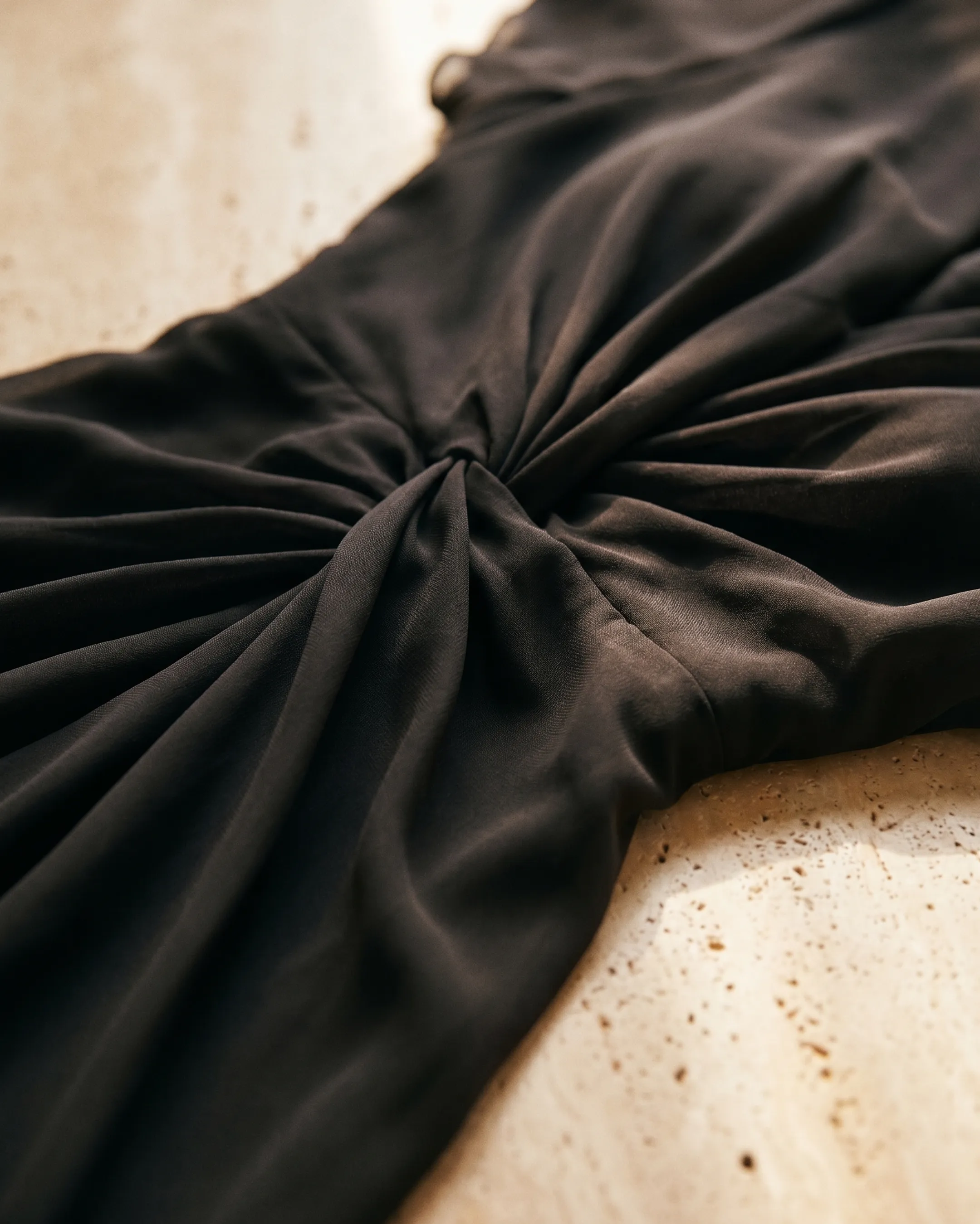 Macro of the Portia dress in Basalt — fluid charcoal chiffon and the centre twist-knot gather on warm travertine, Hugo Caesar Tassi