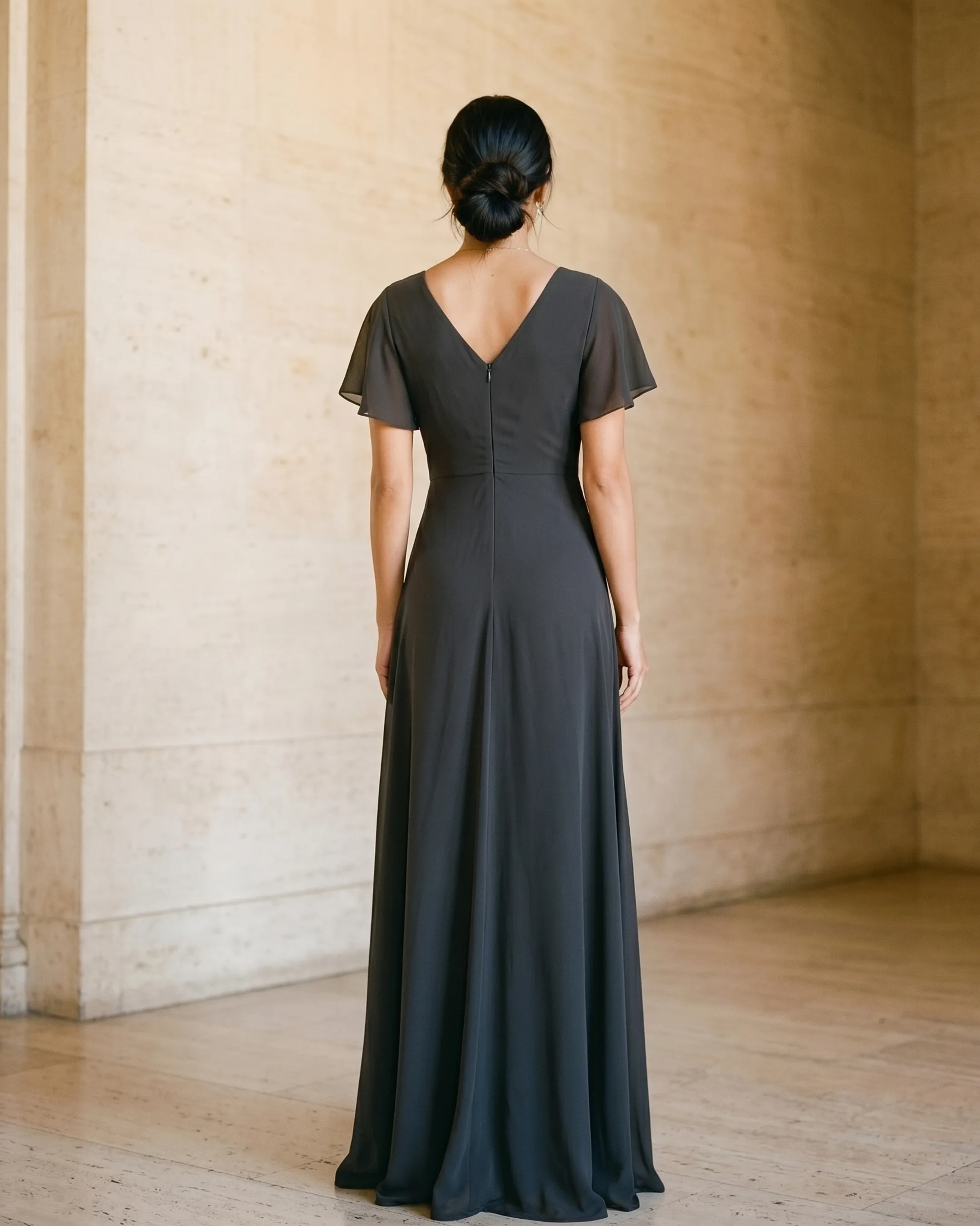 Full-length rear view of the Hugo Caesar Tassi Portia short-sleeve chiffon A-line dress in Basalt, seen from behind, showing the back of the dress, the flutter sleeves, defined twist-knot waist and fluid A-line chiffon skirt in a Roman travertine studio