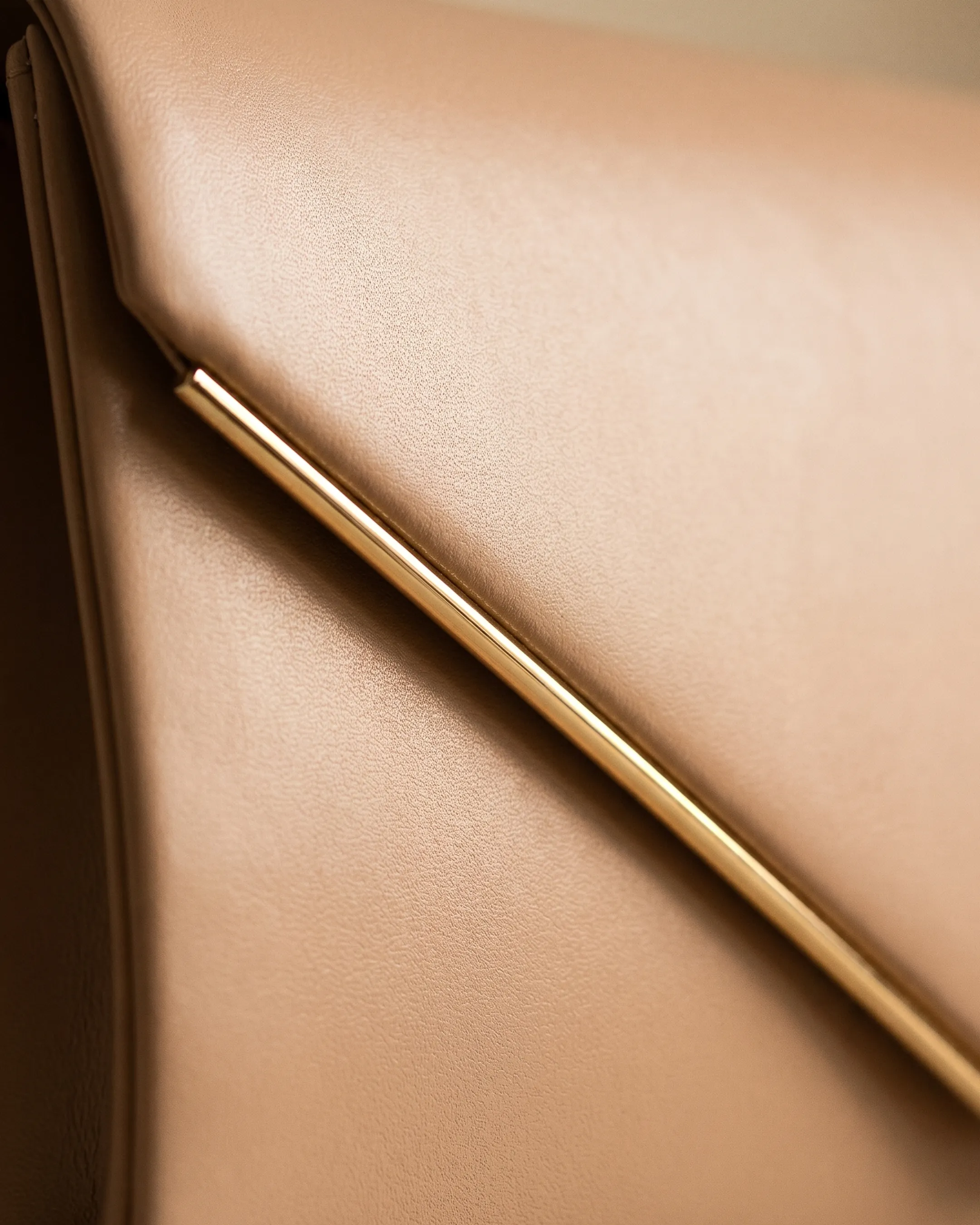 Macro detail of the Hugo Caesar Tassi Quirina smooth nappa leather grain and the fine polished-gold trim line running along the V-shaped envelope flap, in Travertine camel