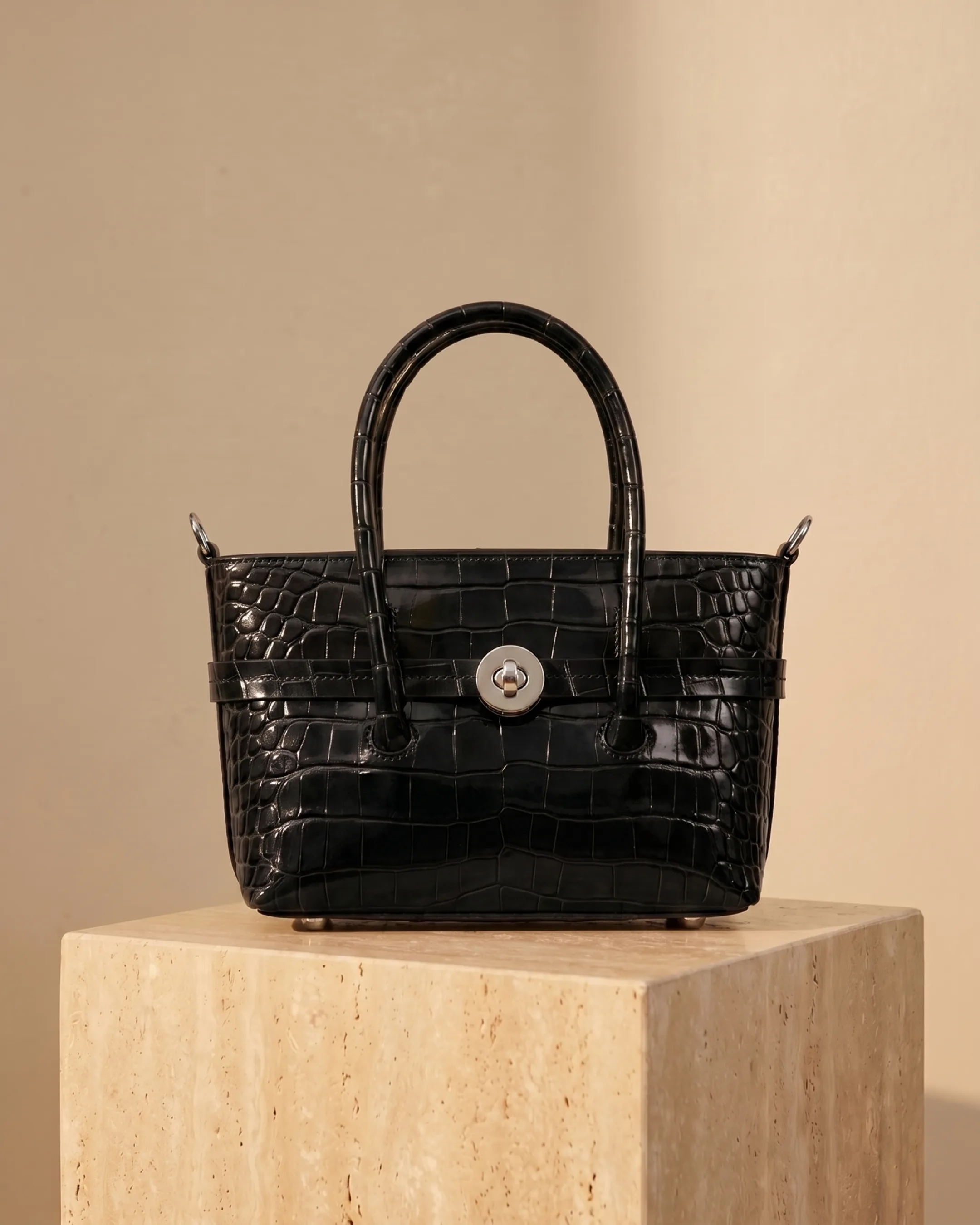 Hugo Caesar Tassi Salaria structured top-handle satchel in Basalt black croc-embossed leather with silver turn-lock, on a warm travertine pedestal