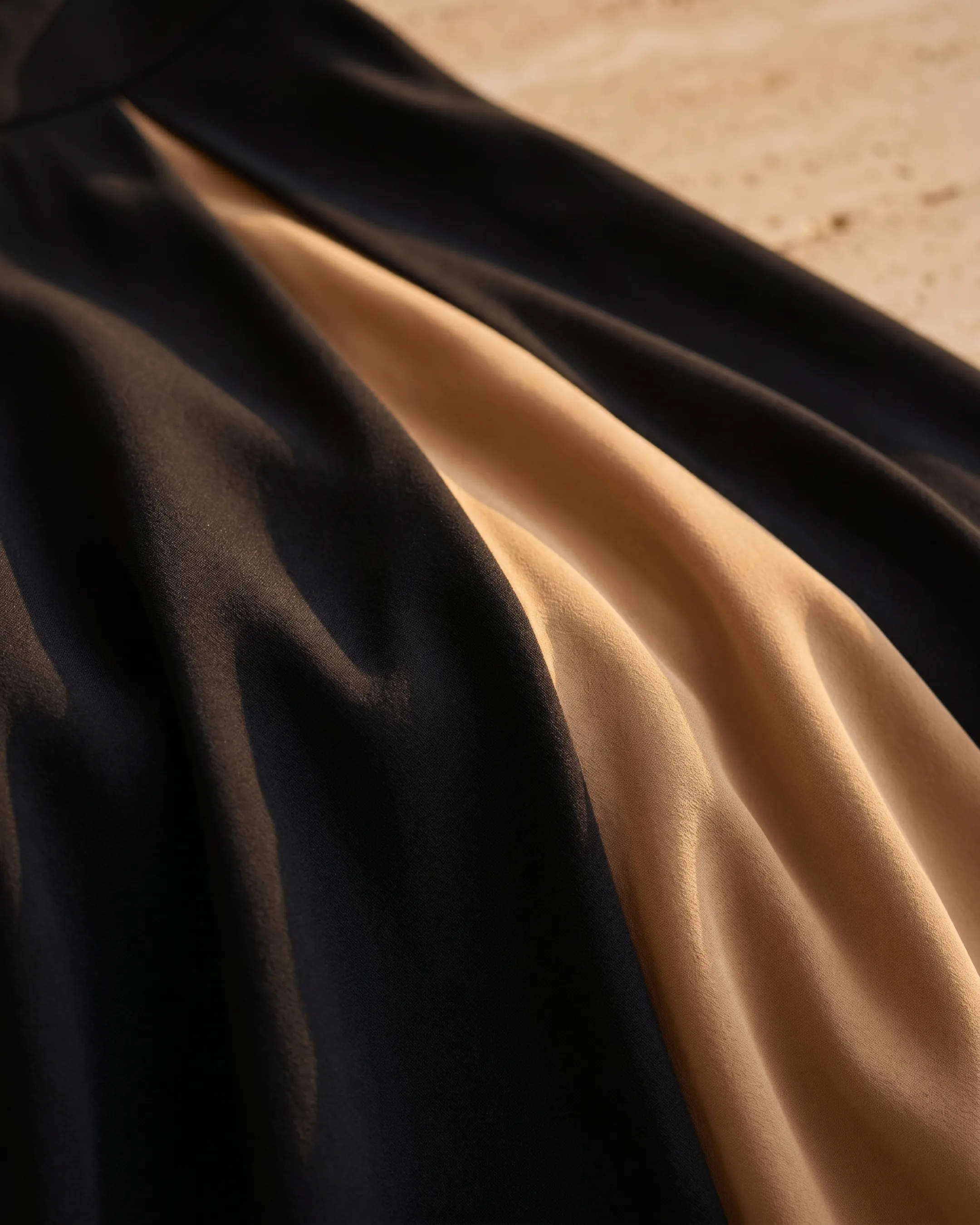Hugo Caesar Tassi Sentia macro of the fluid matte crepe where deep Basalt black meets the warm Travertine-tan godet, soft folds catching golden light