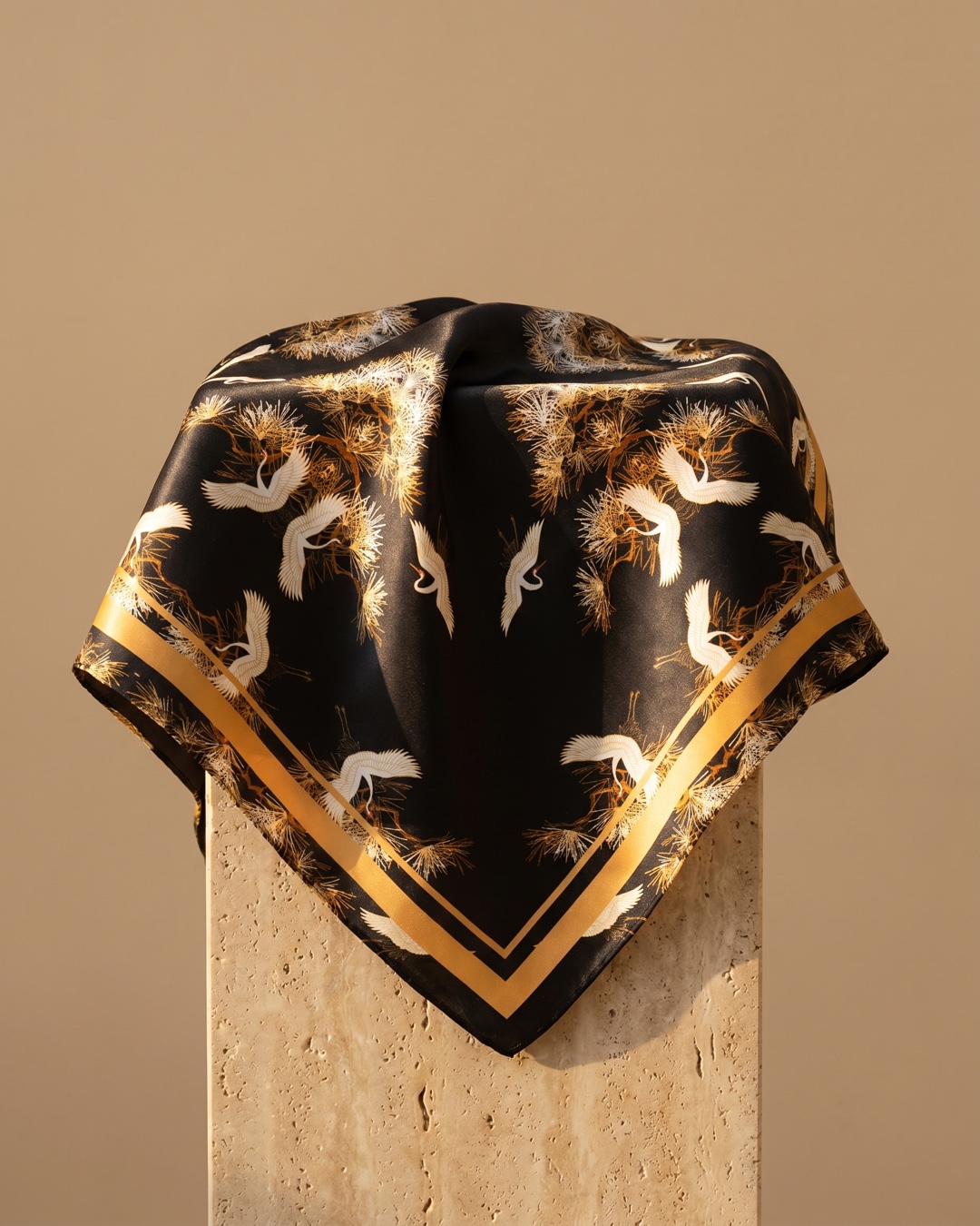 Hugo Caesar Tassi Spoleto silk square scarf in black crepe-satin with white flying cranes, golden pine boughs and a gold border, draped on a warm travertine pedestal