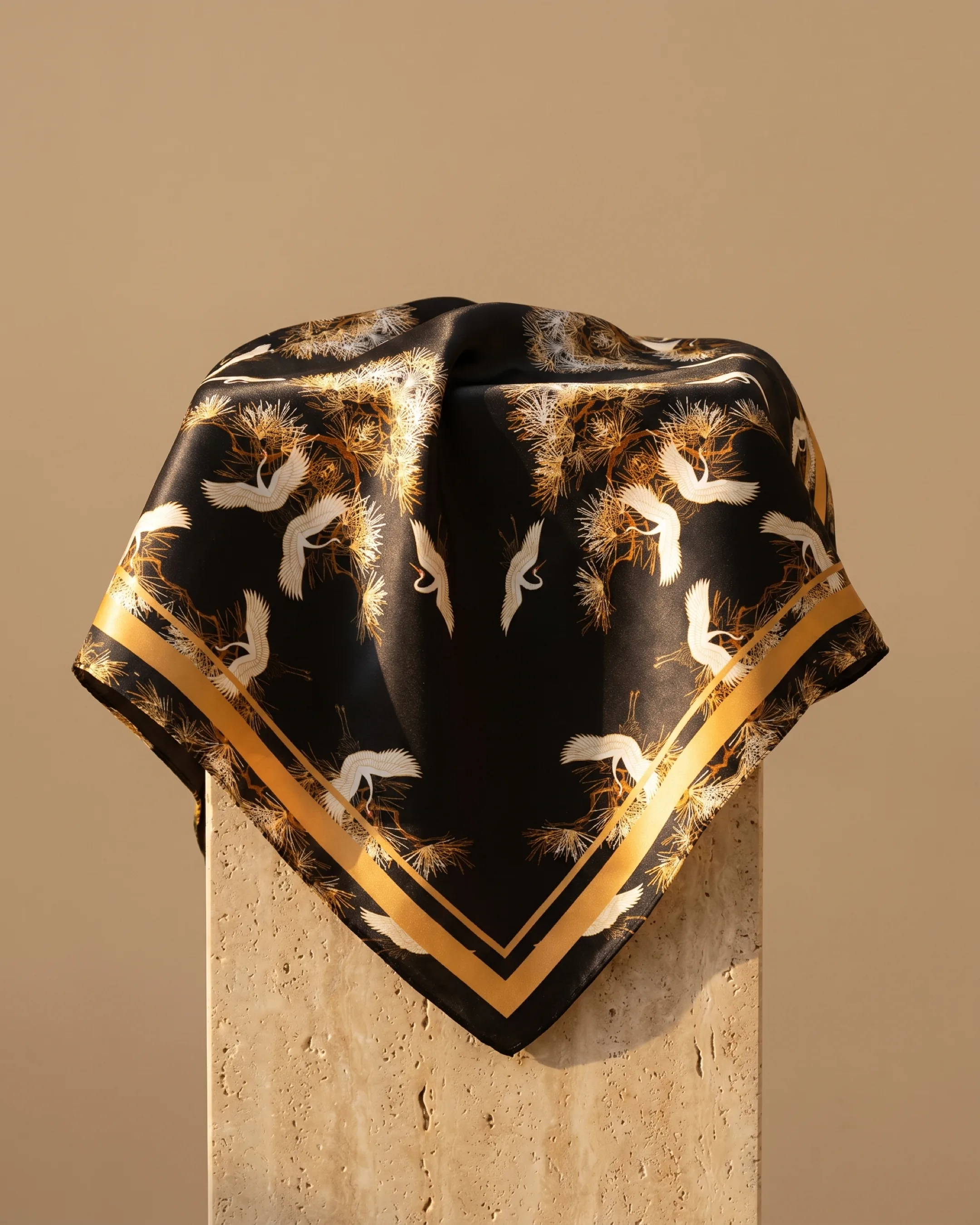 Hugo Caesar Tassi Spoleto silk square scarf in black crepe-satin with white flying cranes, golden pine boughs and a gold border, draped on a warm travertine pedestal