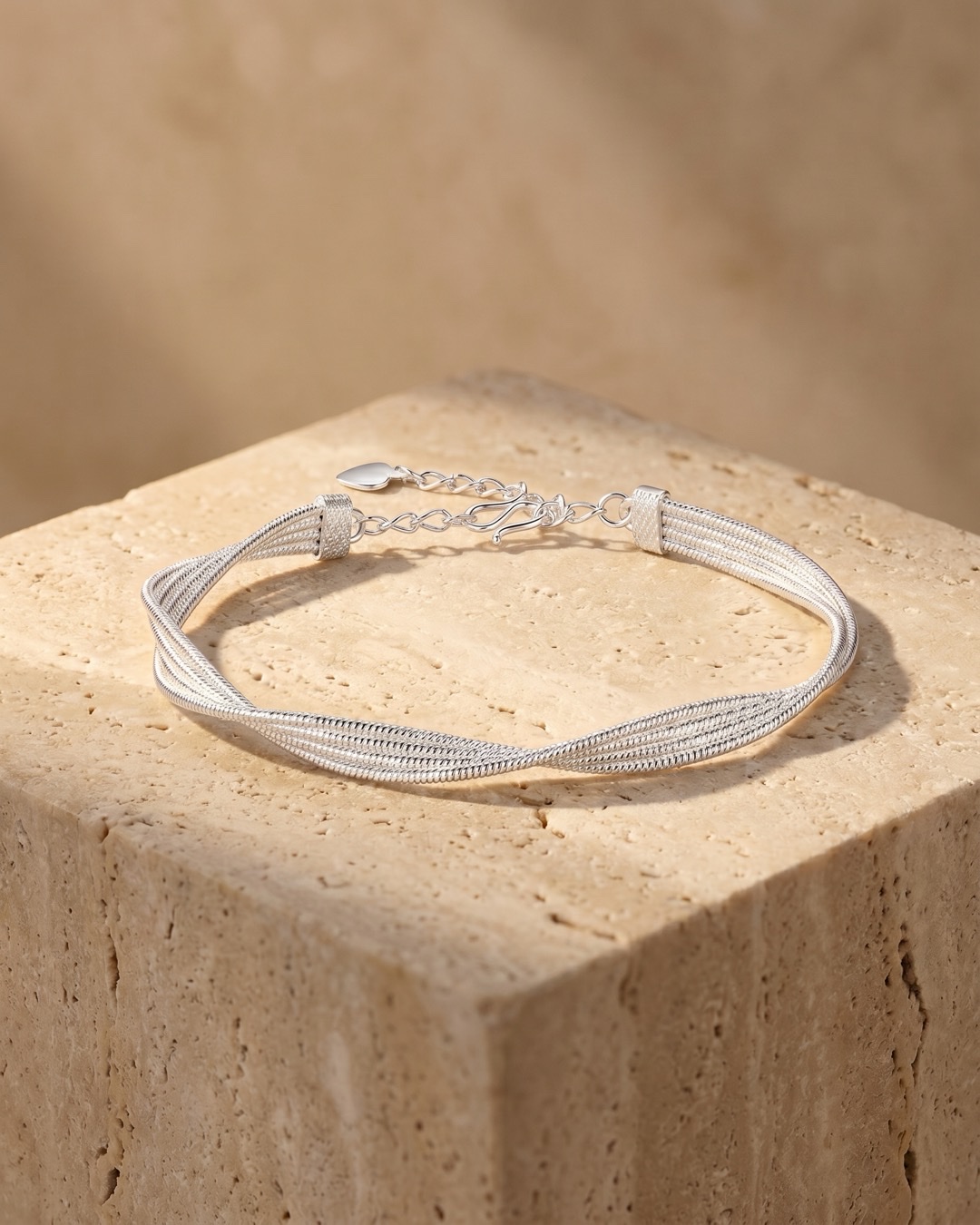Hugo Caesar Tassi Stella twisted multi-strand fine silver mesh bracelet with a heart charm and lobster clasp, resting on a warm travertine pedestal