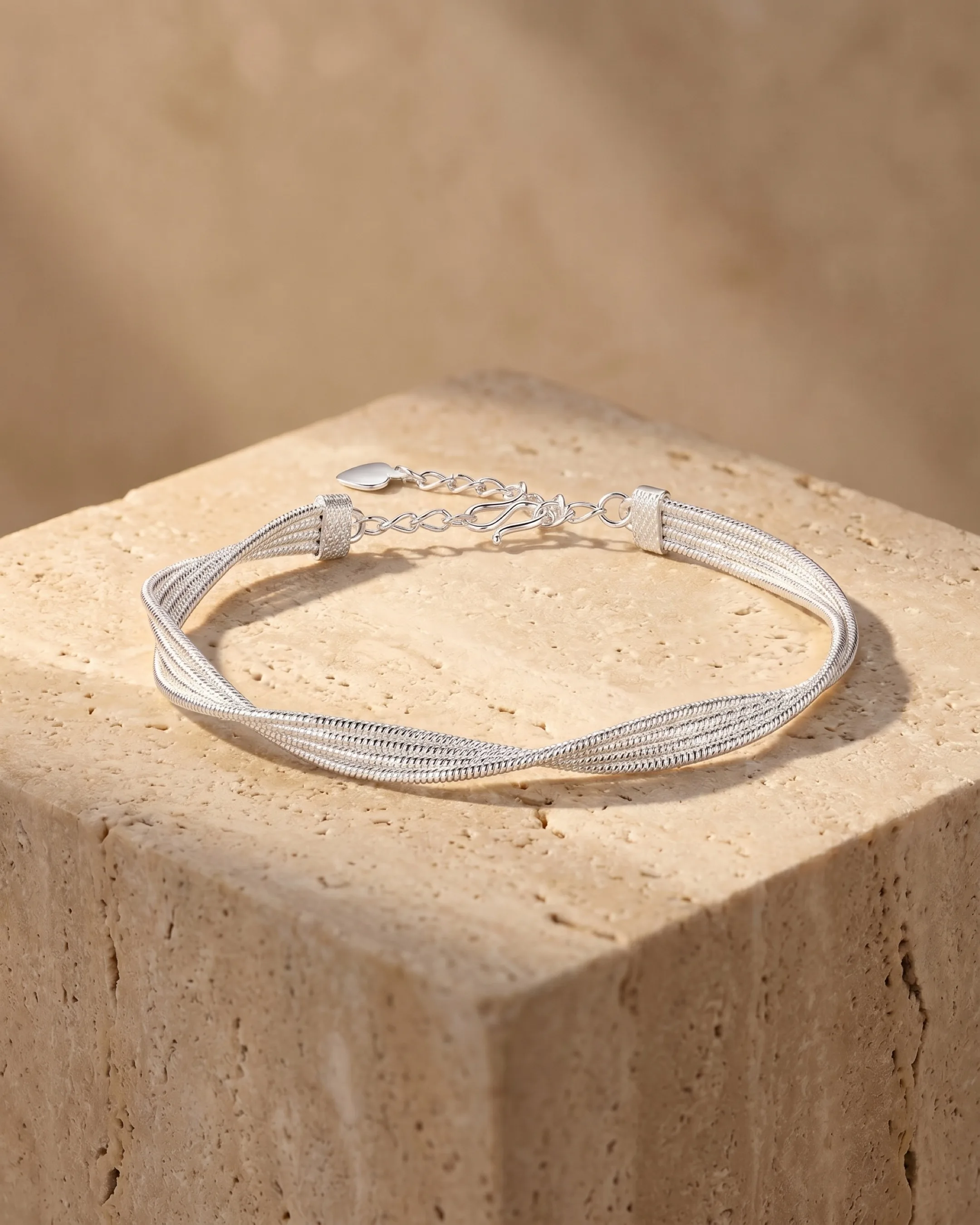Hugo Caesar Tassi Stella twisted multi-strand fine silver mesh bracelet with a heart charm and lobster clasp, resting on a warm travertine pedestal
