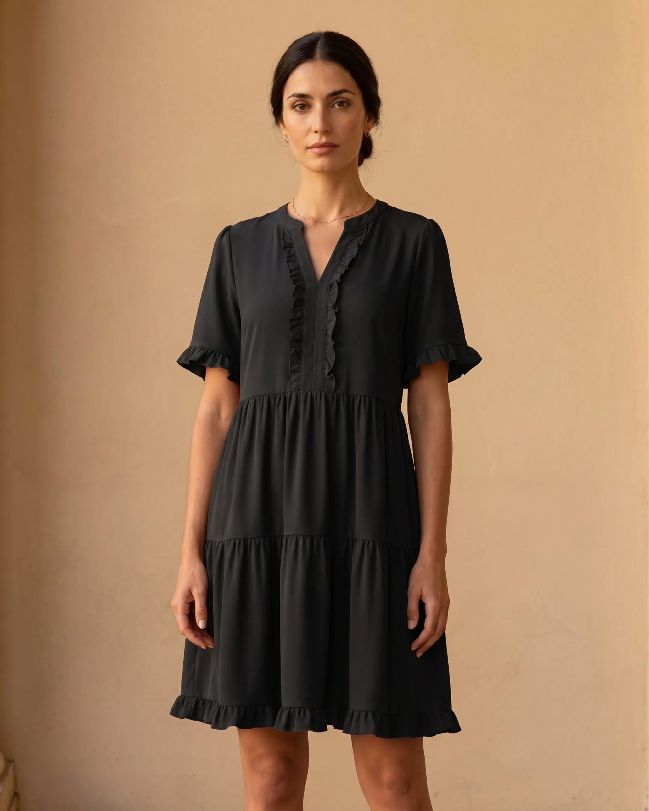 Hugo Caesar Tassi Tertulla split V-neck ruffle-trim tiered swing dress in Basalt black, full-length front view by a travertine column
