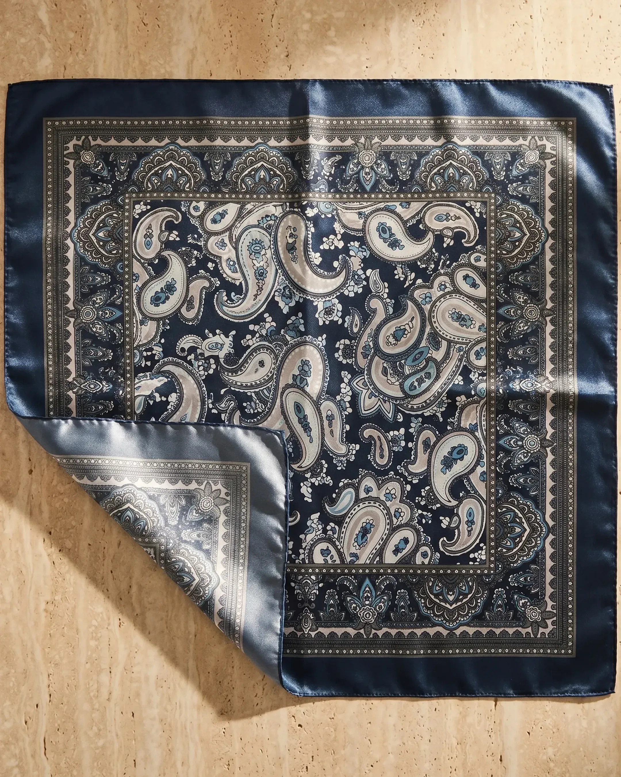 The Hugo Caesar Tassi Tessera scarf in Cobalto styled flat on warm travertine, showing the full navy-and-cream paisley print and folded satin corner
