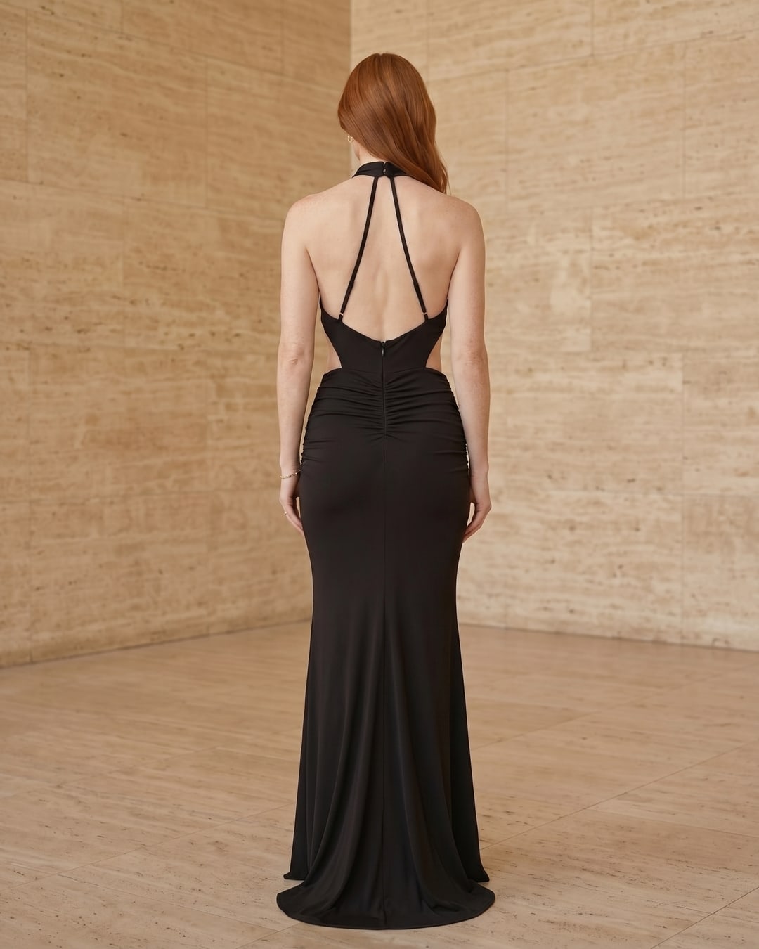 Full-length rear view of the Hugo Caesar Tassi Tullia pleated column evening gown in Basalt black crepe, seen from behind, showing the back of the dress, the open back, halter tie at the nape and fine knife-pleated column sweeping the floor in a Roman travertine studio