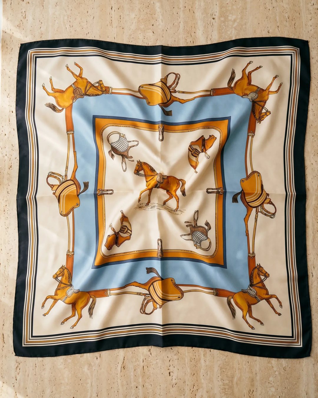 Hugo Caesar Tassi Vesta silk-look square scarf in Avorio shown open as a flat-lay on warm travertine marble, revealing the full equestrian carré print of amber horses and saddlery framed in navy