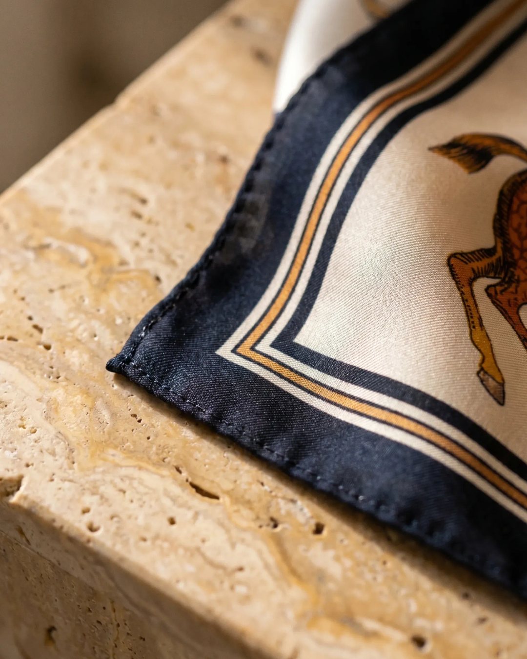 Macro detail of the Hugo Caesar Tassi Vesta scarf hand-rolled twill edge and silk-look weave in Avorio ivory with golden-amber and navy print