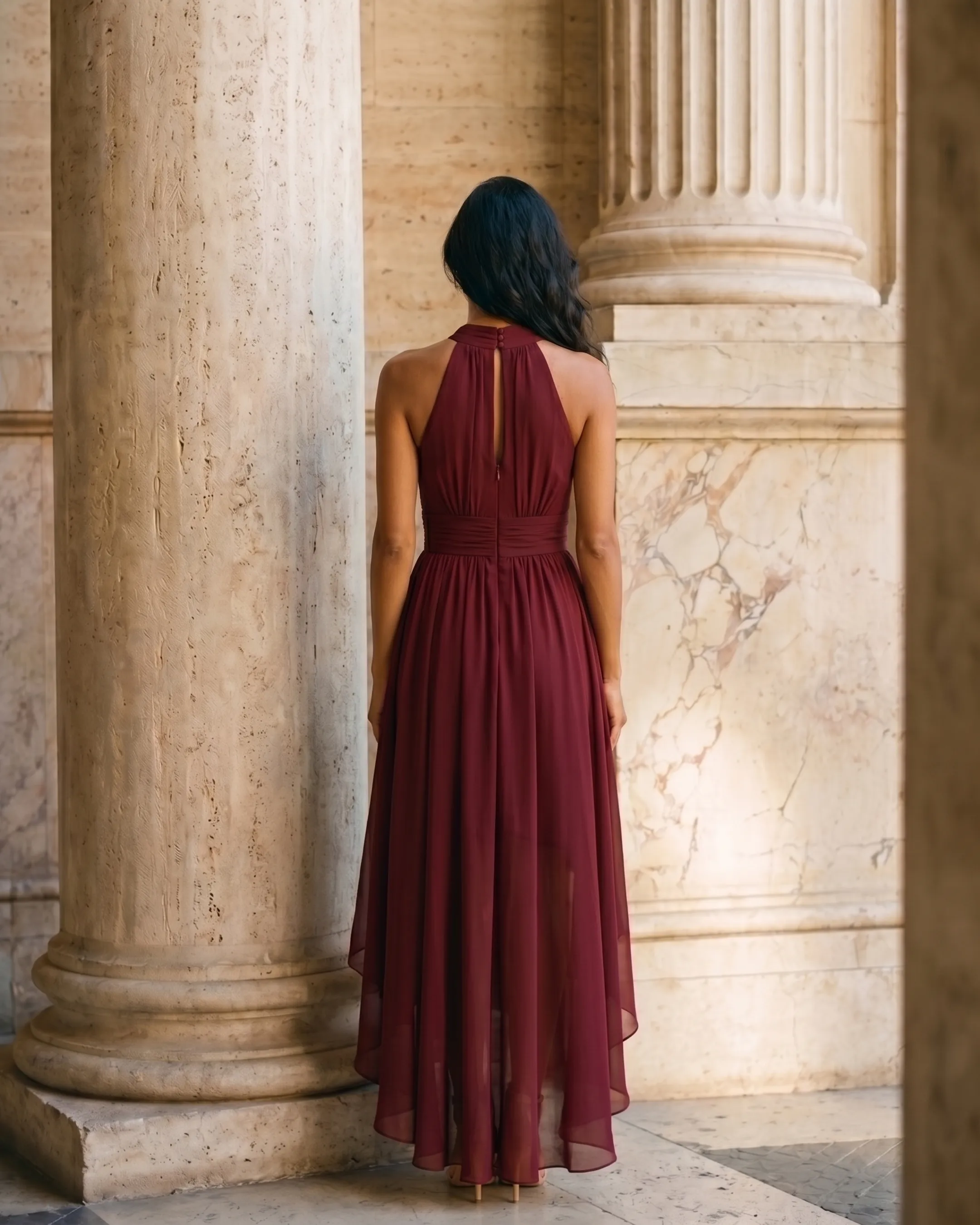 Full-length rear view of the Hugo Caesar Tassi Vibia halter-neck chiffon evening dress in deep wine-red Porpora, seen from behind, showing the back of the dress, the halter tie at the nape, open back and sweeping high-low chiffon hem in a Roman travertine studio