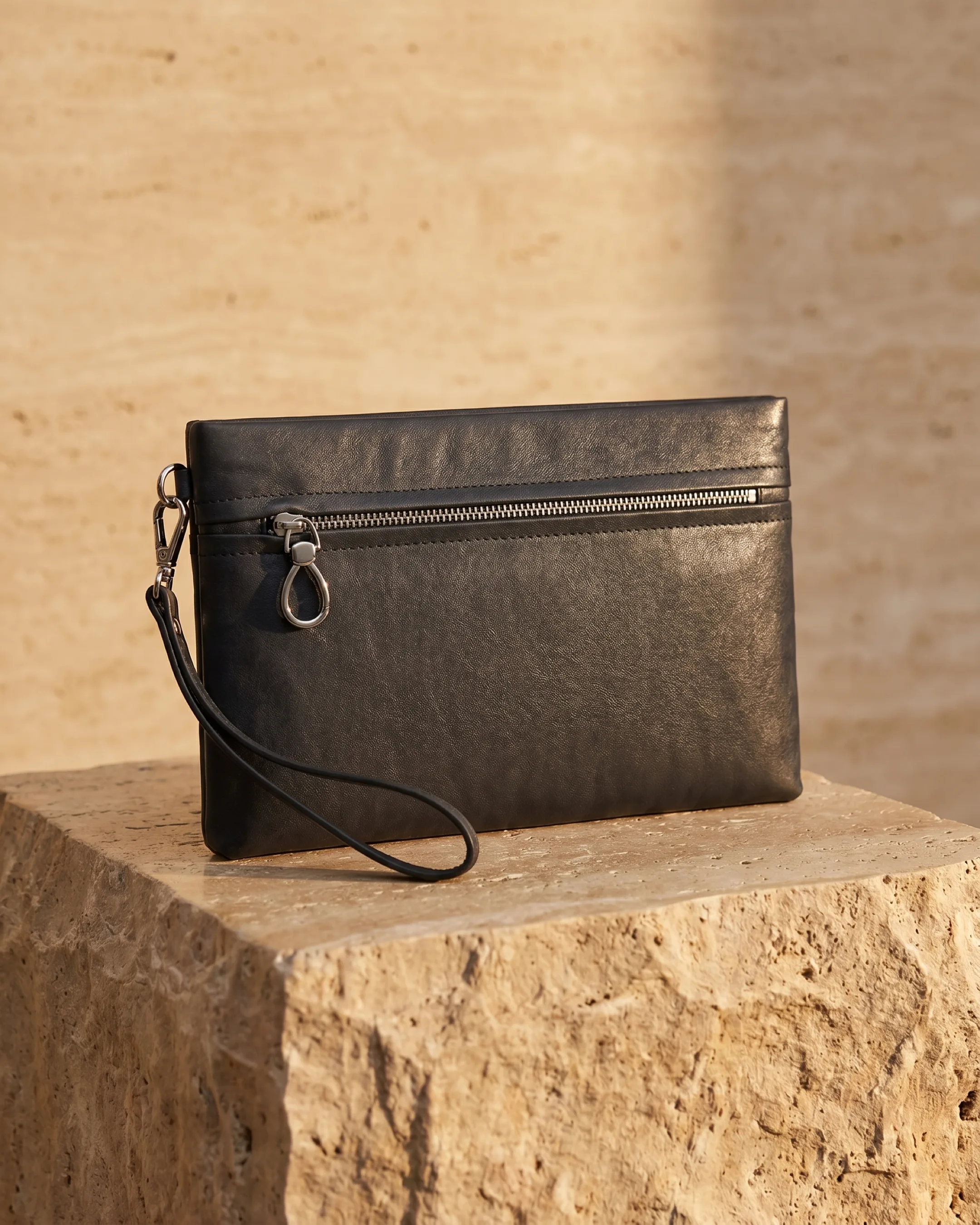 Hugo Caesar Tassi Viminia small leather evening clutch in Basalt black, top-zip with silver pull and wrist strap, on a warm travertine pedestal